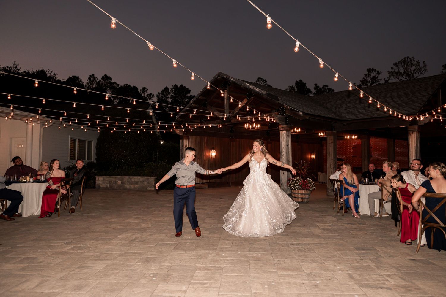 String lights illuminate an outdoor evening wedding reception dance floor as people dance and celebrate.