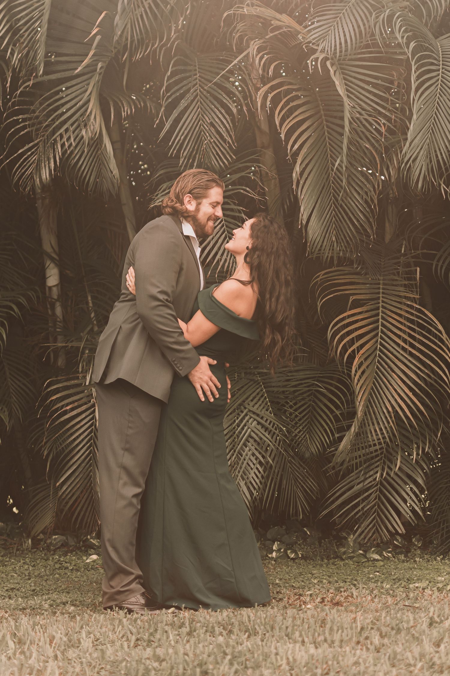 A romantic couple in formal attire embrace in front of a tropical palm leaf backdrop during a special occasion.