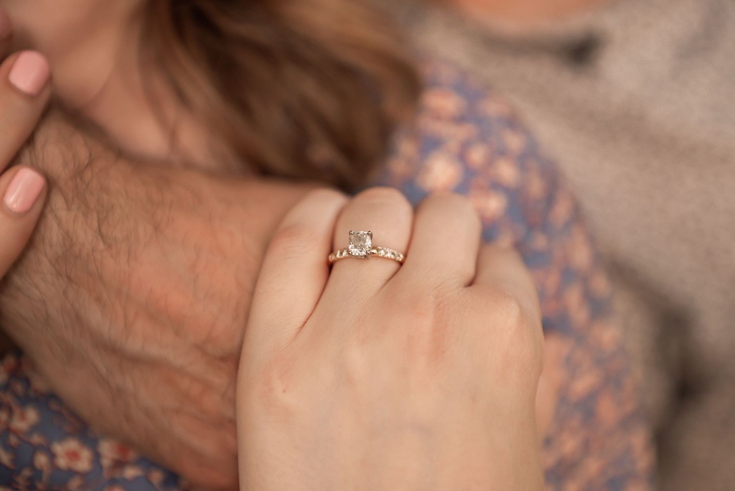 Close up view of an engagement ring being worn on a hand against a floral patterned background.