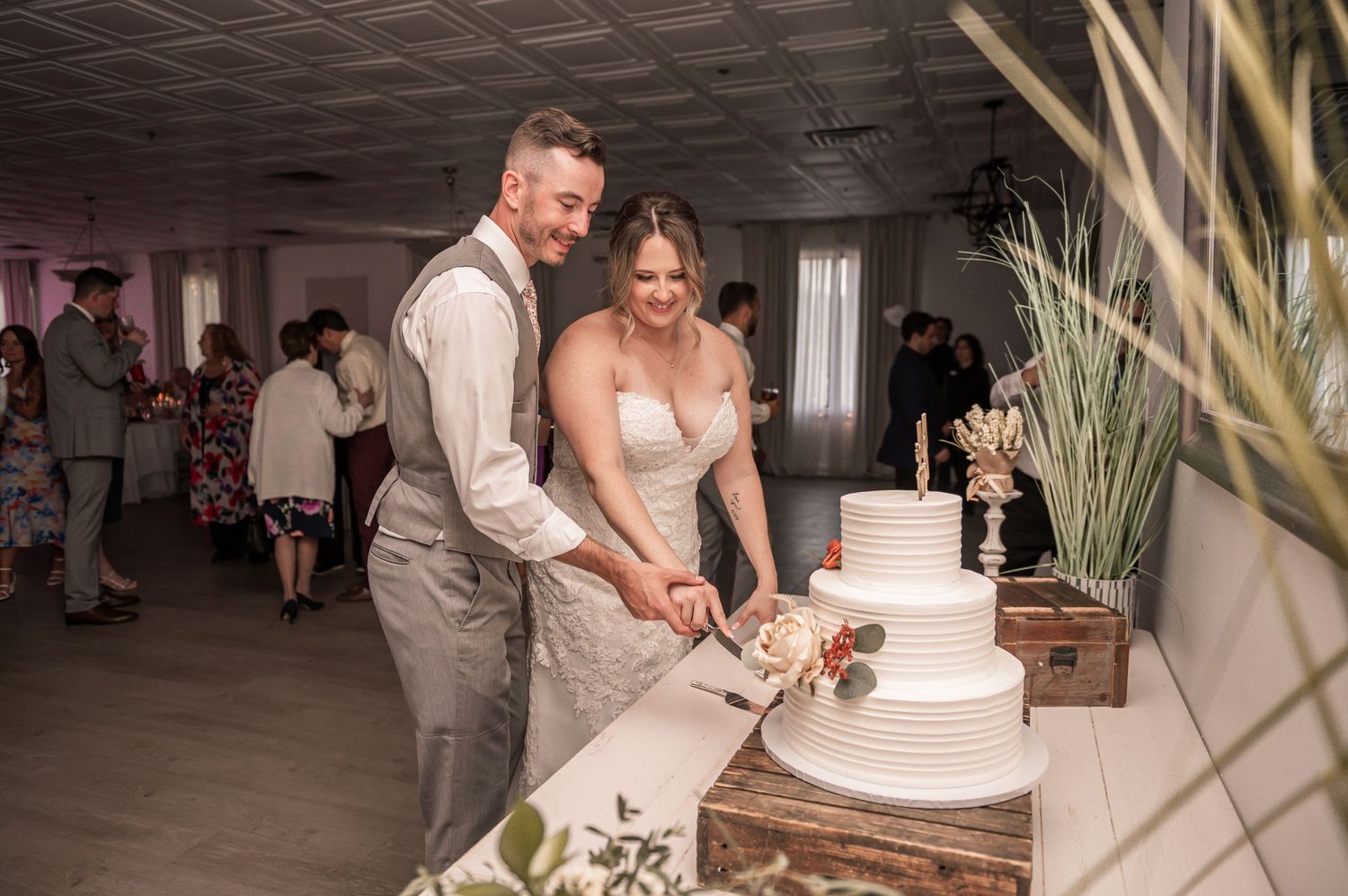 Wedding reception with a tall white cake displayed as couples dance in an indoor venue with decorative plants.