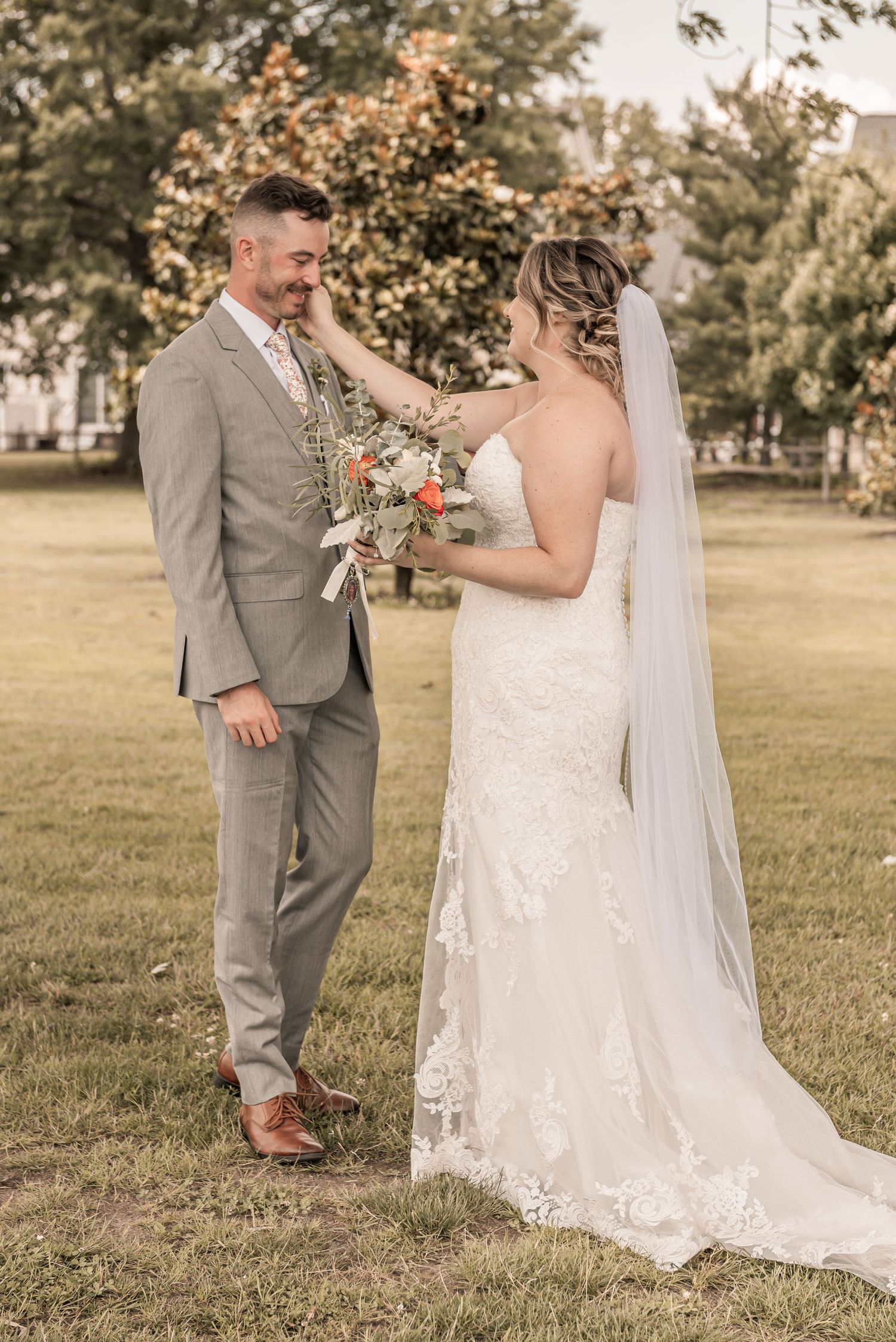 A romantic outdoor wedding scene with a couple in elegant gray suit and lace gown walking together in a sunlit field.