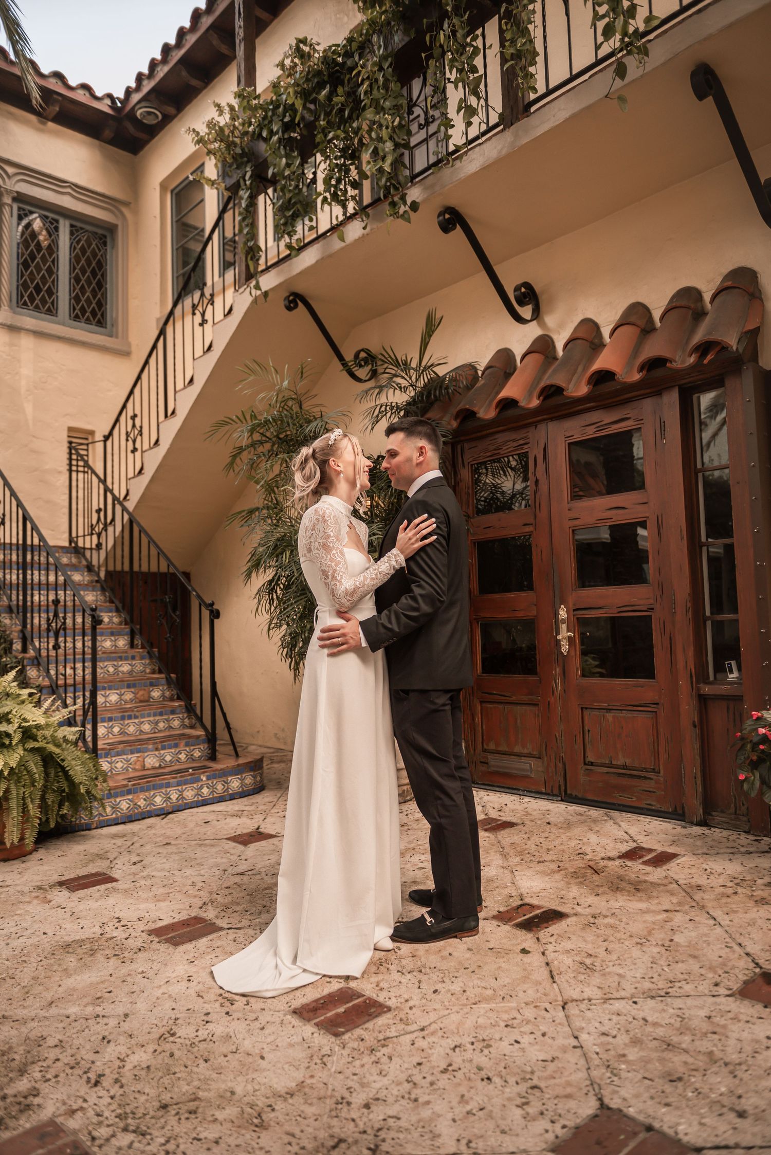 Bride and groom embrace in a kiss outside a Spanish-style villa courtyard with wrought iron staircase.