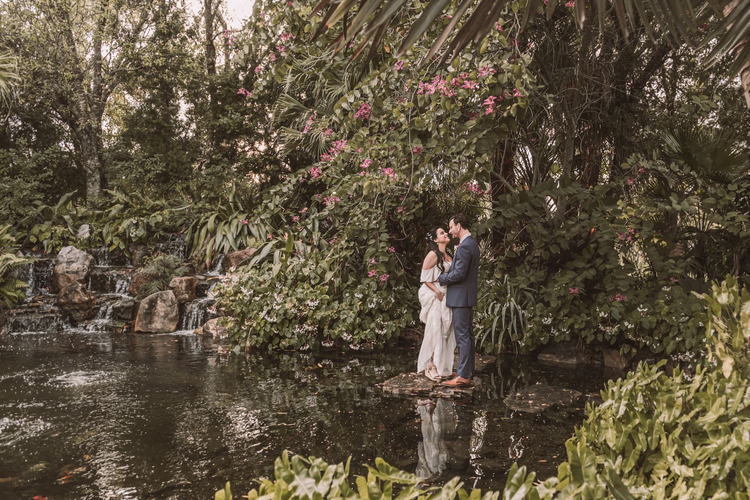 A romantic scene unfolds at a garden pond with flowering trees and reflections in the water.
