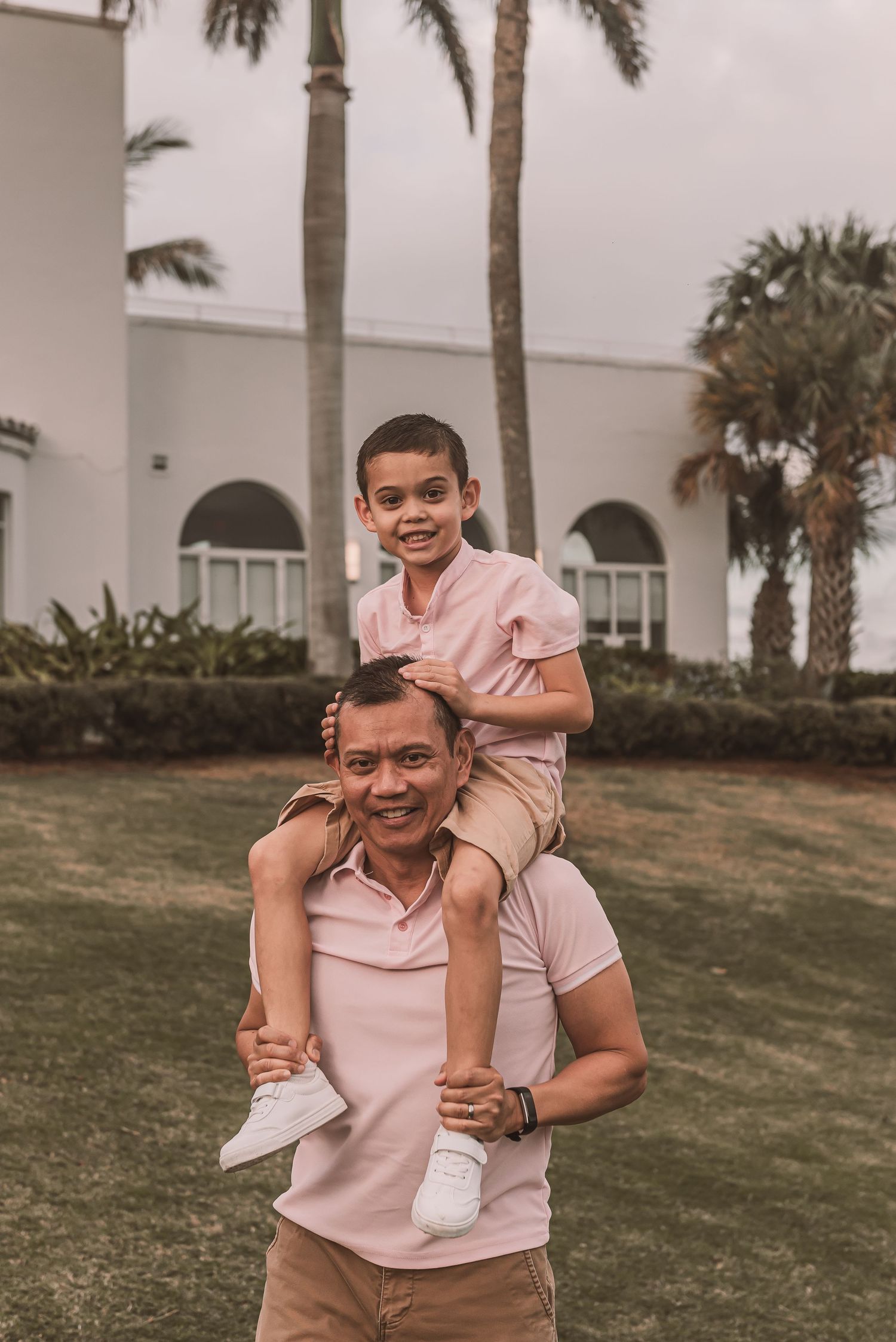 Family members wearing matching pink polo shirts share a playful moment outdoors with palm trees and arched building in background.