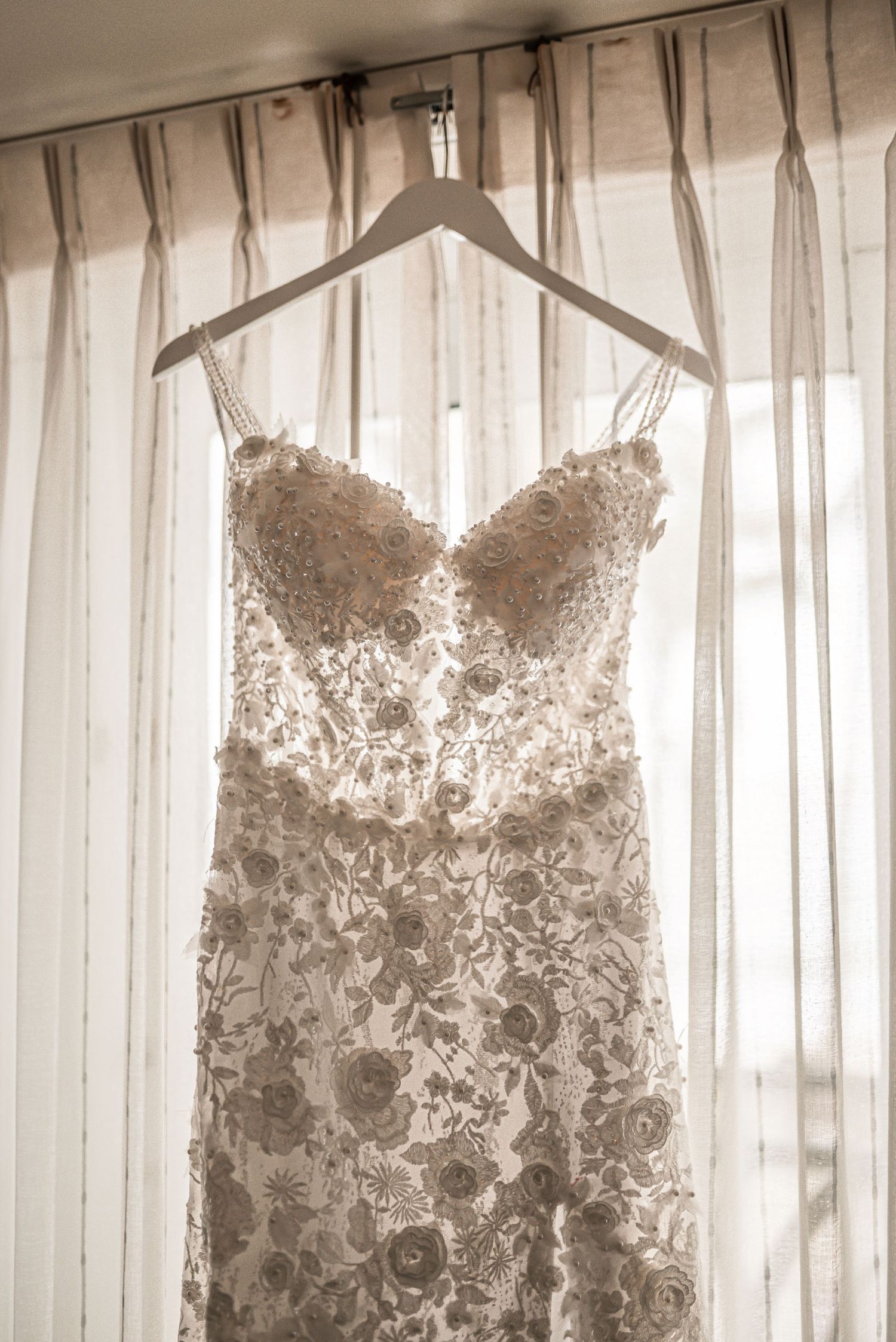 Three elegant lace wedding dresses with intricate floral patterns hang from hangers against a white curtained background.