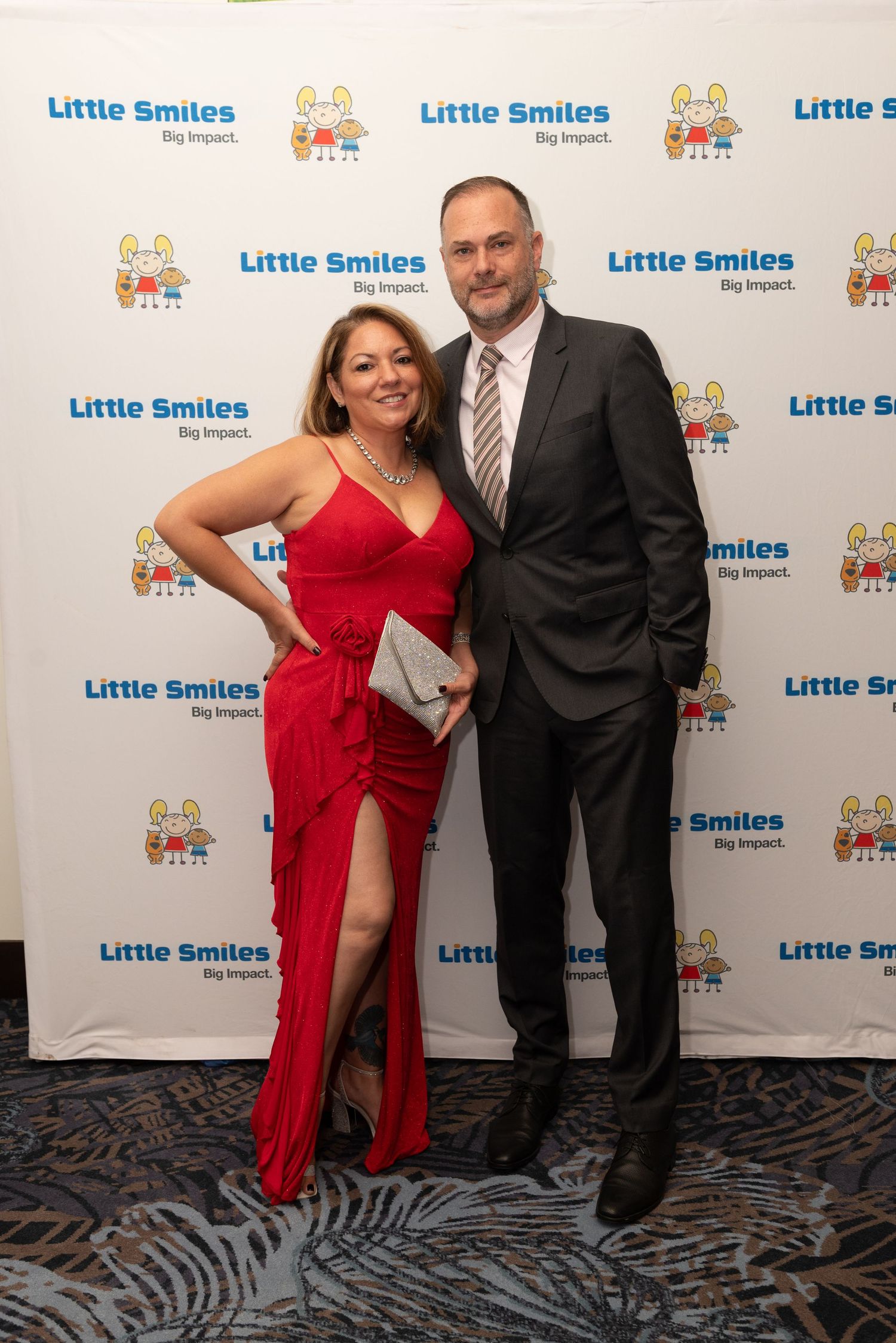 A couple poses for photos at a Little Smiles event backdrop, with the woman wearing a red dress.
