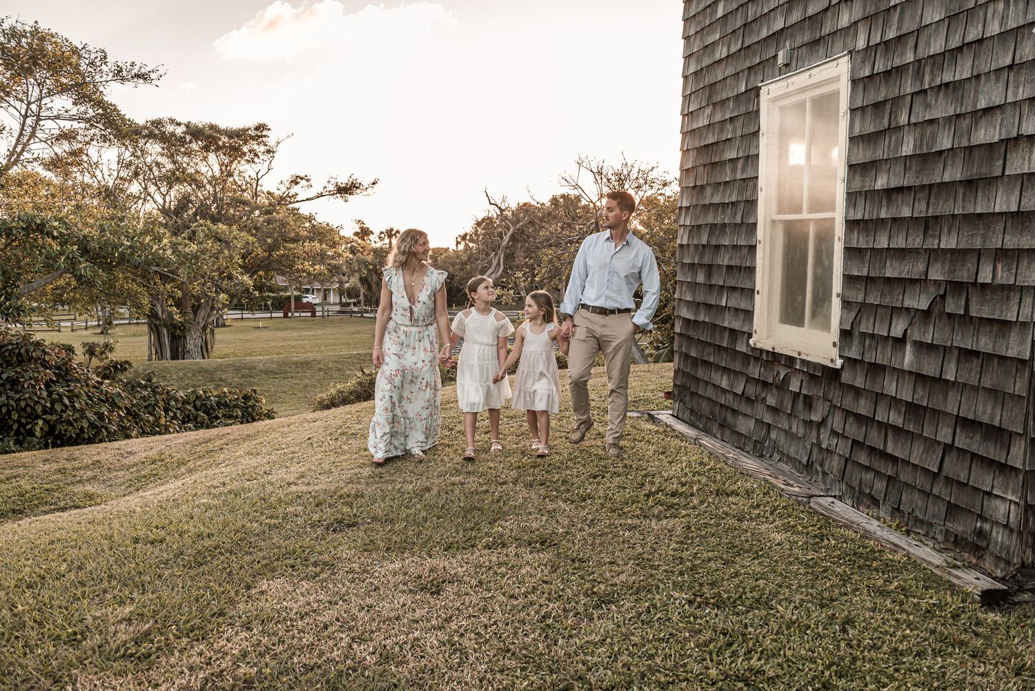 A group in light summer attire walks together on a grassy slope beside a shingled house at sunset.