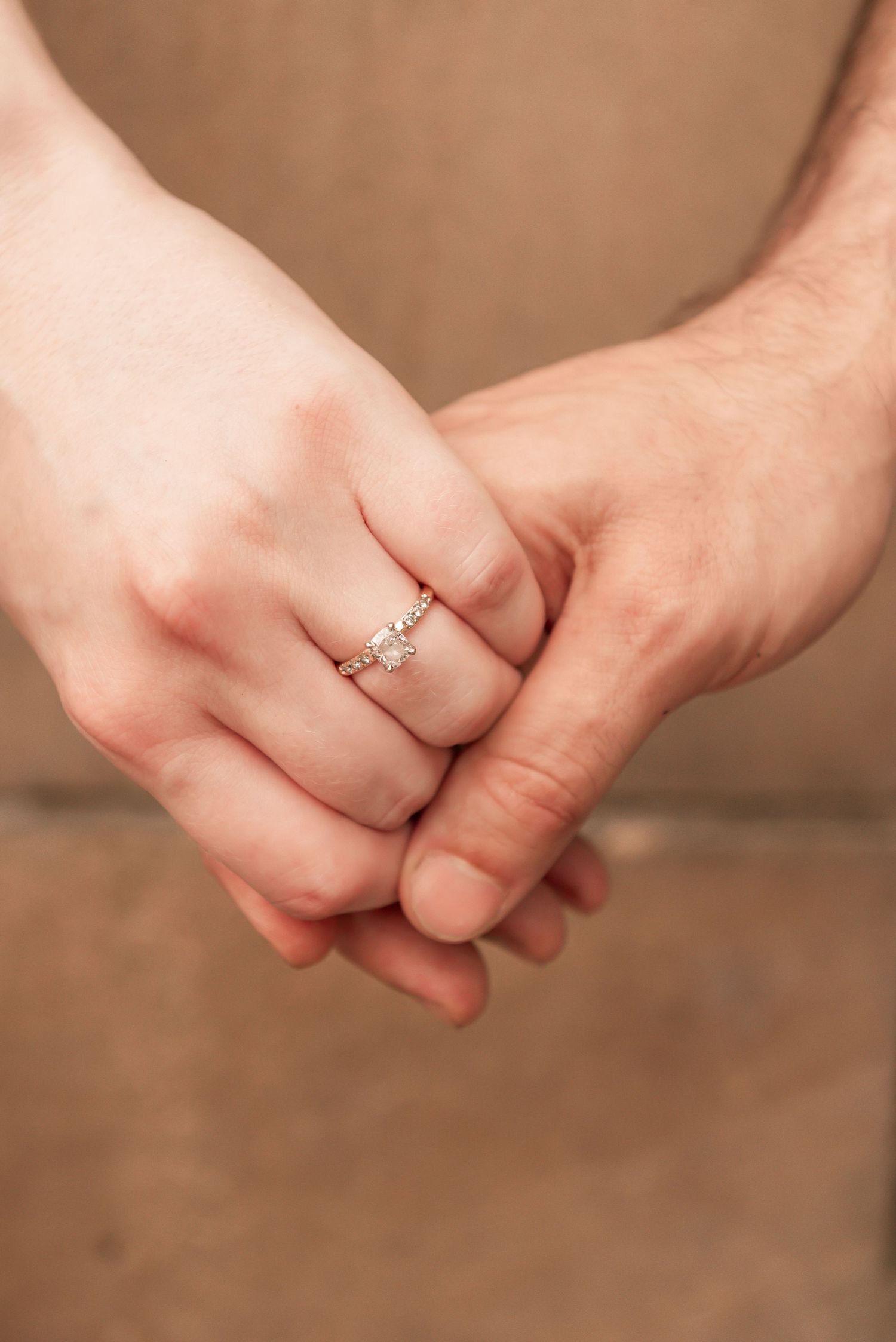 Close-up photo of clasped hands showing an elegant solitaire engagement ring against a neutral background.