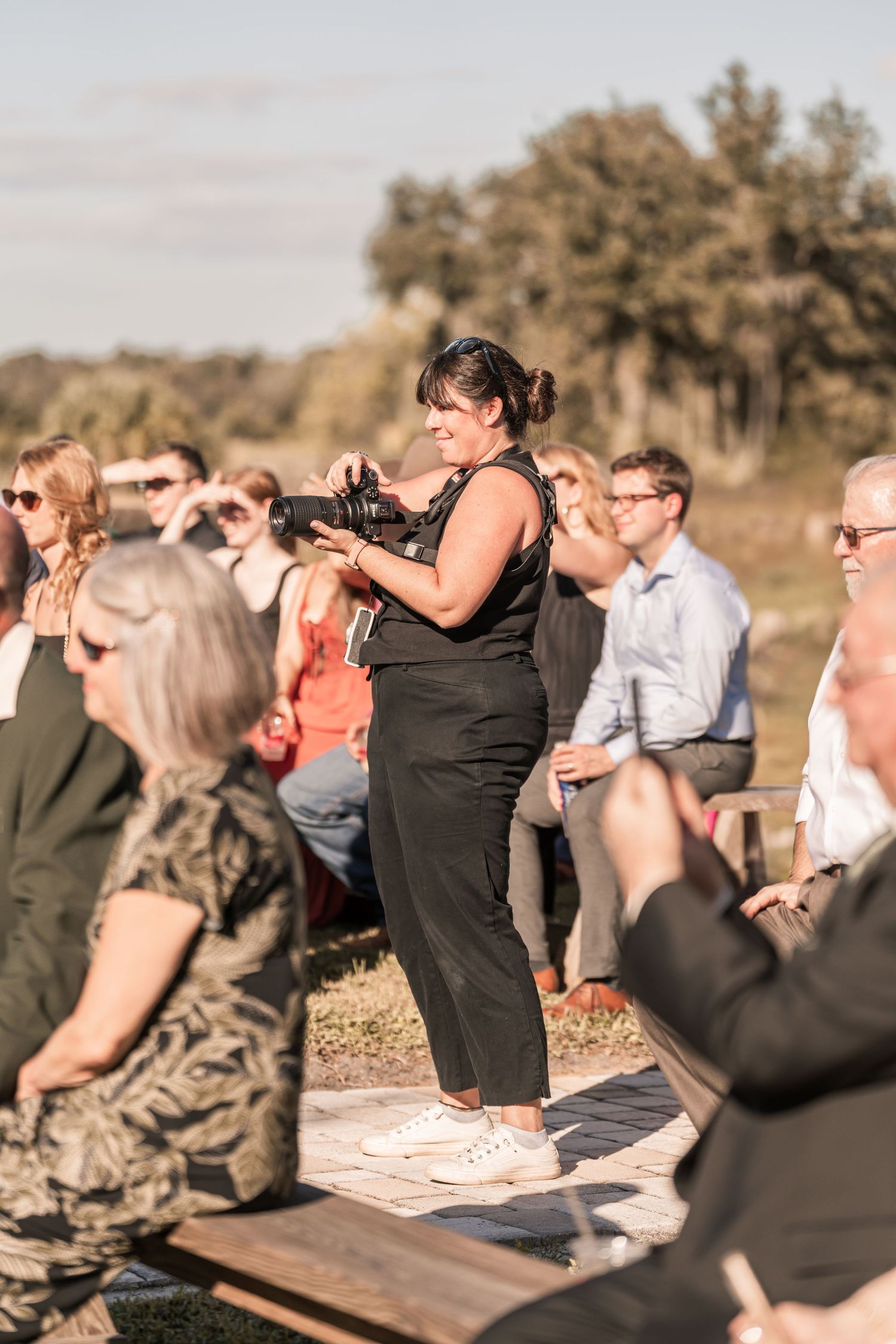 Photographer in black outfit capturing moments at an outdoor event with guests seated in sunny field.