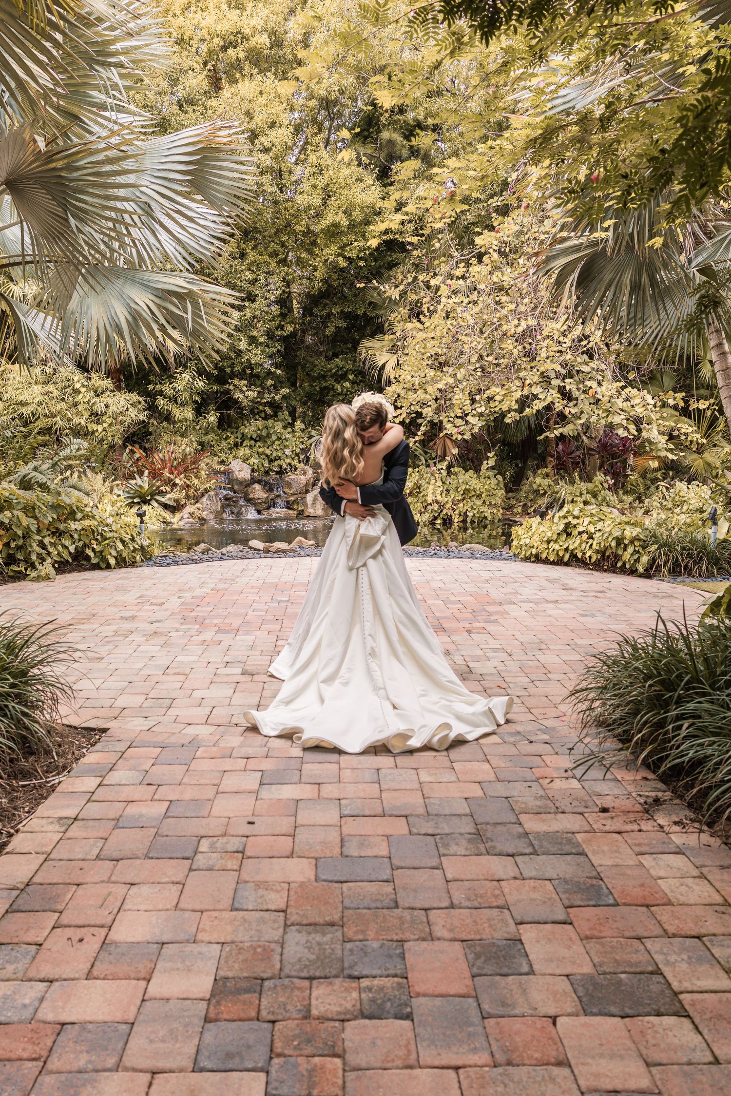 Bride and groom embracing on a brick path surrounded by lush tropical gardens on their wedding day.