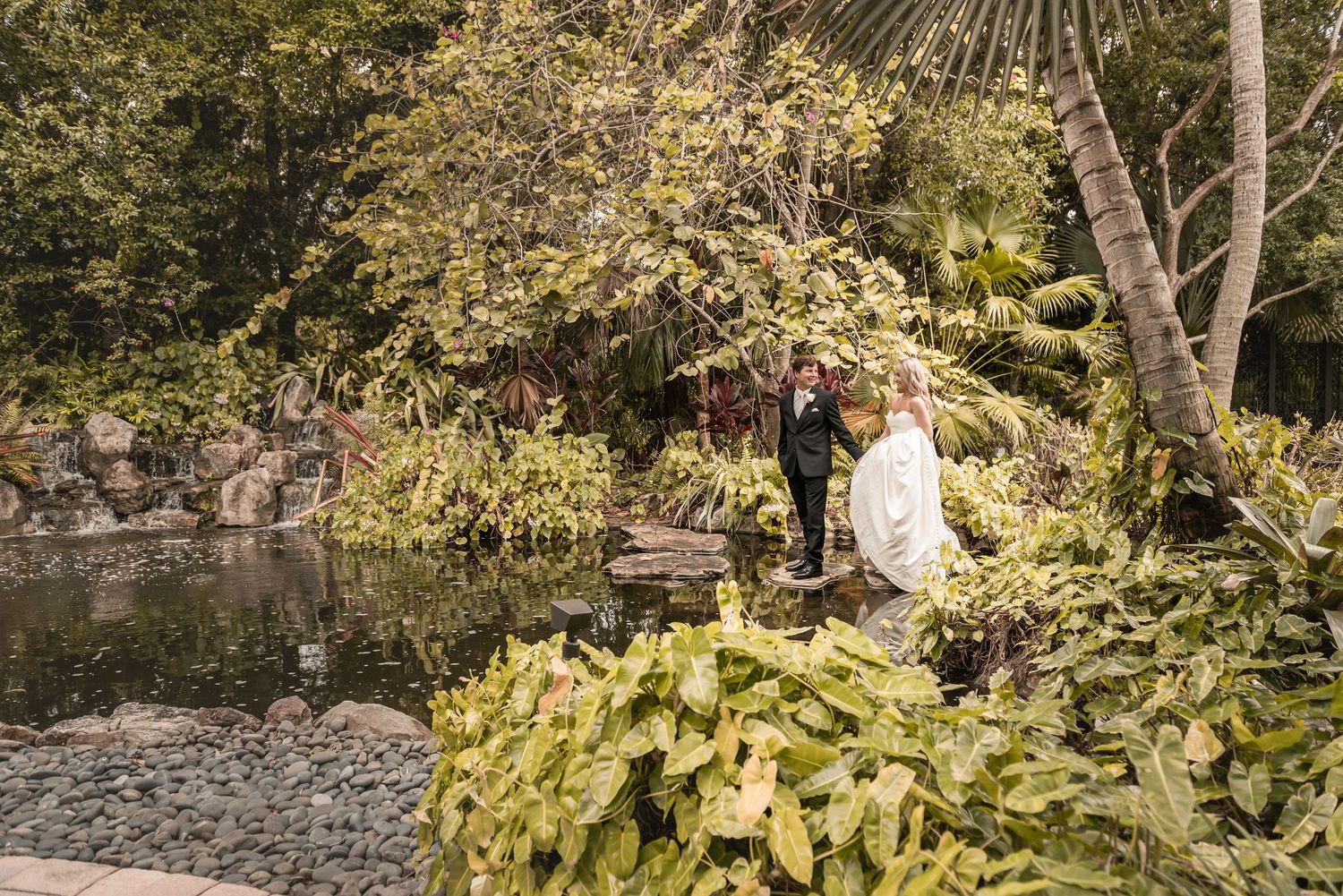 Bride and groom standing by a serene pond surrounded by lush tropical greenery in an outdoor garden wedding setting.