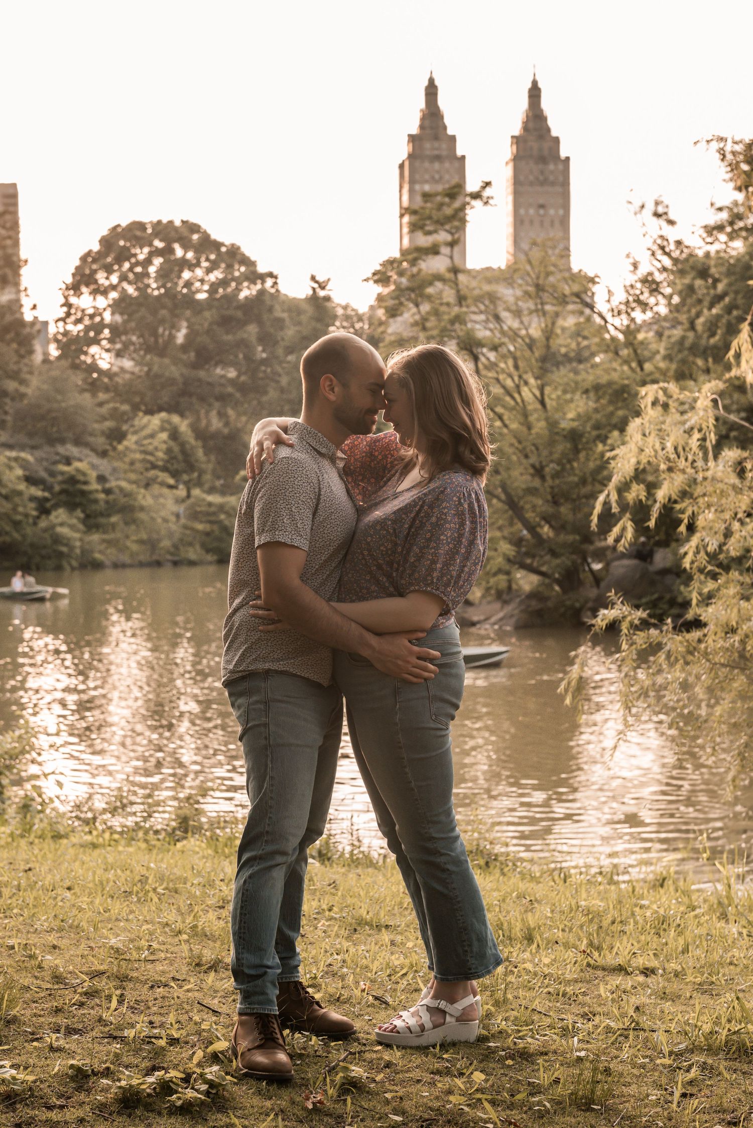 A romantic couple embraces near a lake in Central Park with the iconic twin towers of the San Remo building visible at sunset.