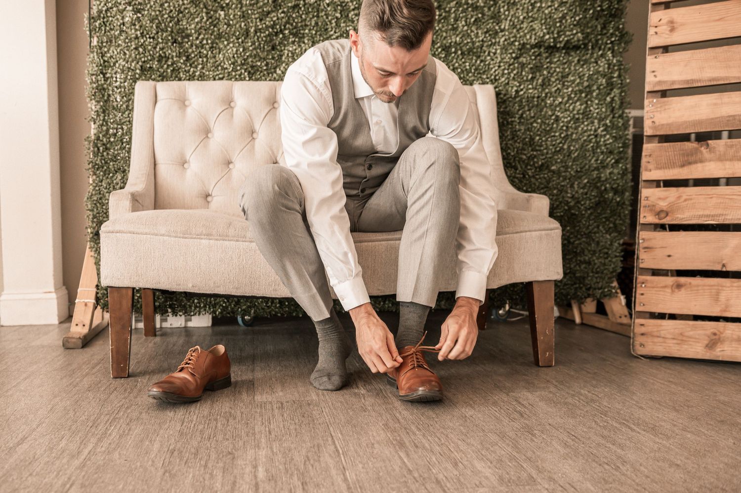 Groom tying brown leather dress shoes while seated in elegant rustic setting with tufted chairs and wooden decor.
