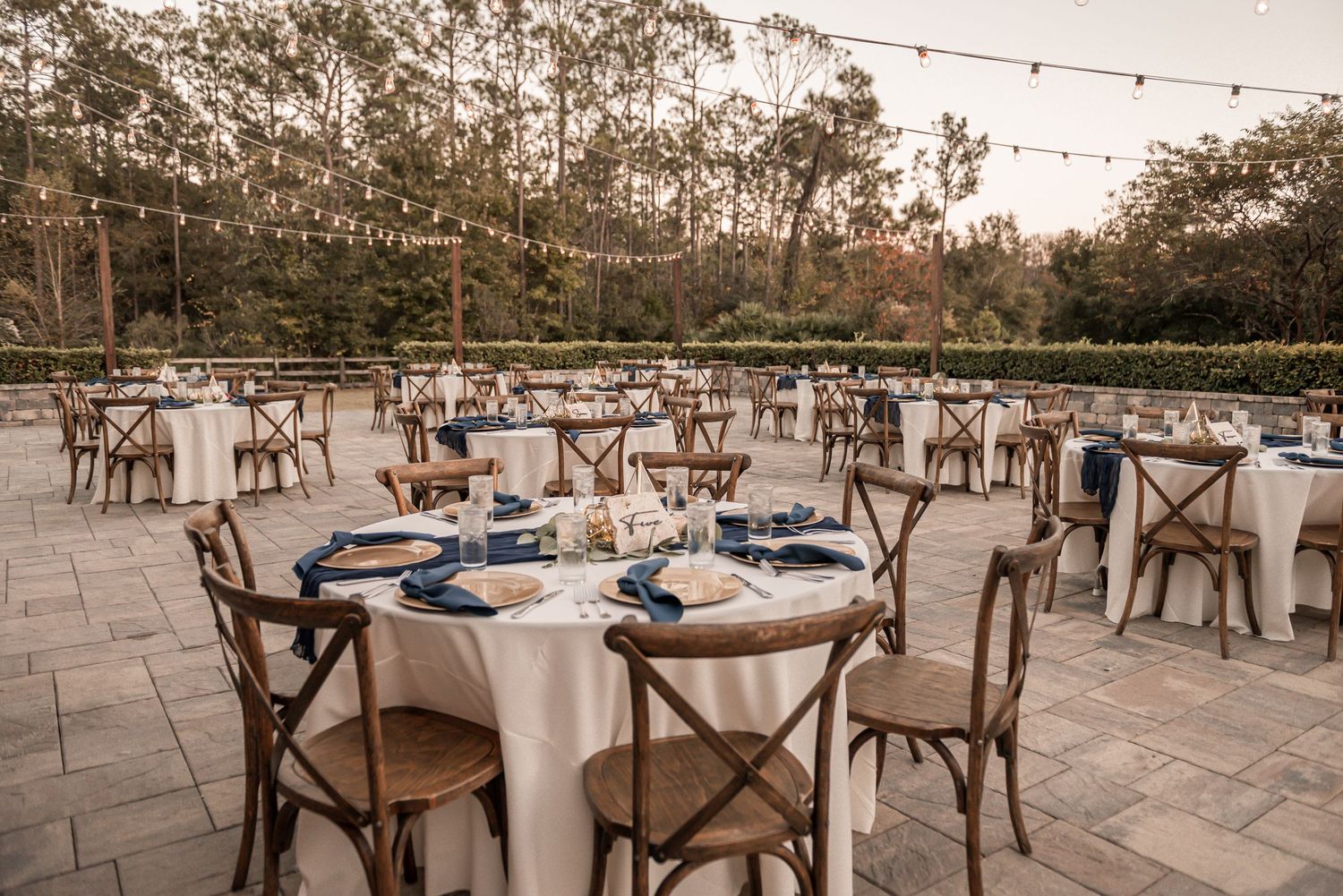 Outdoor wedding reception setup with round tables, white linens and wooden cross-back chairs on a stone patio at dusk.