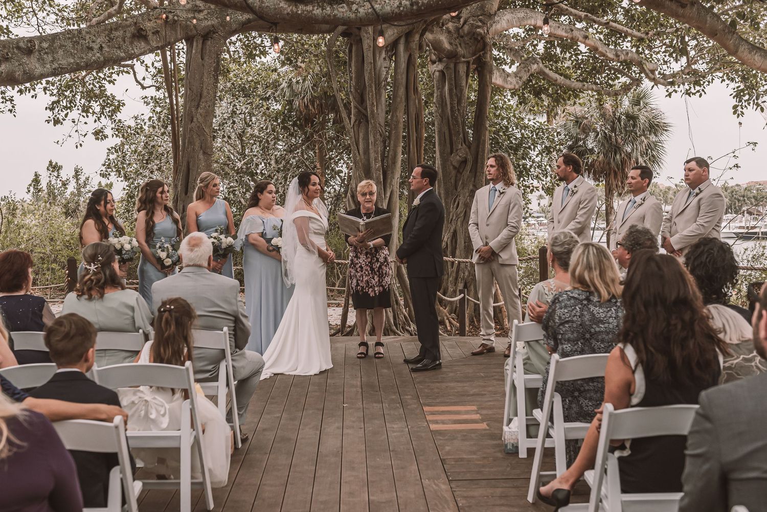 Wedding guests seated in white chairs on a rustic outdoor deck surrounded by trees during a ceremony.