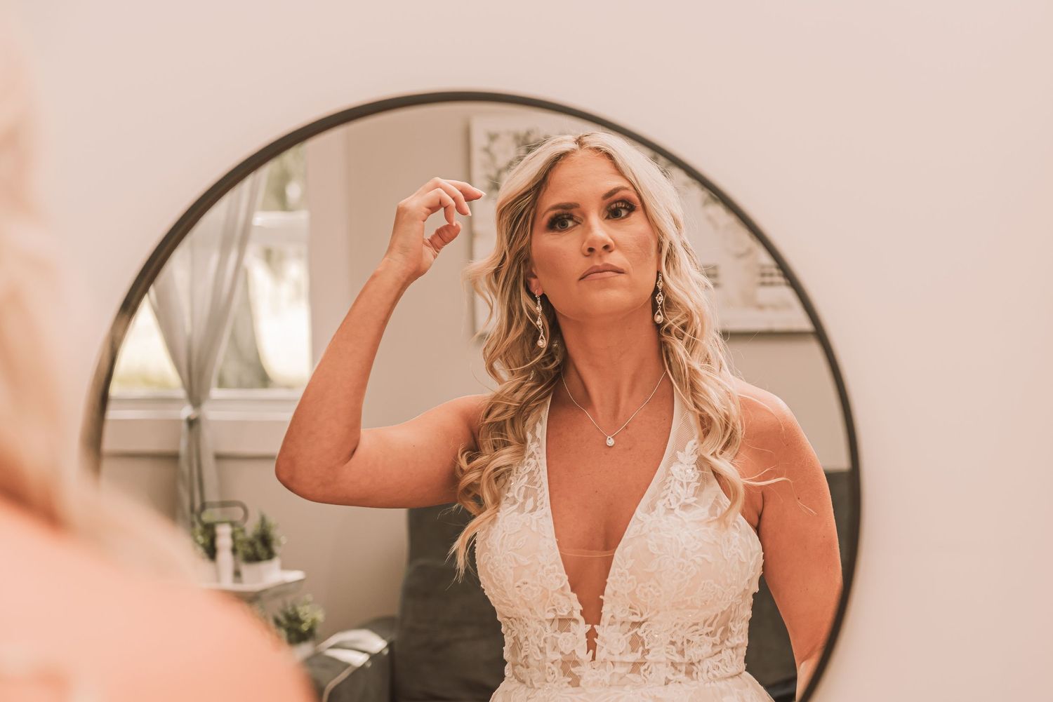 A reflection in a round mirror shows someone in a white lace wedding dress adjusting their long blonde curled hair.
