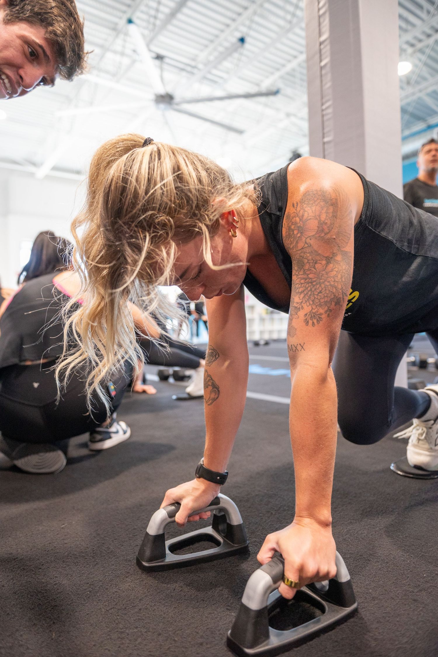 Tattooed woman doing push-ups on handles during intense gym workout competition.