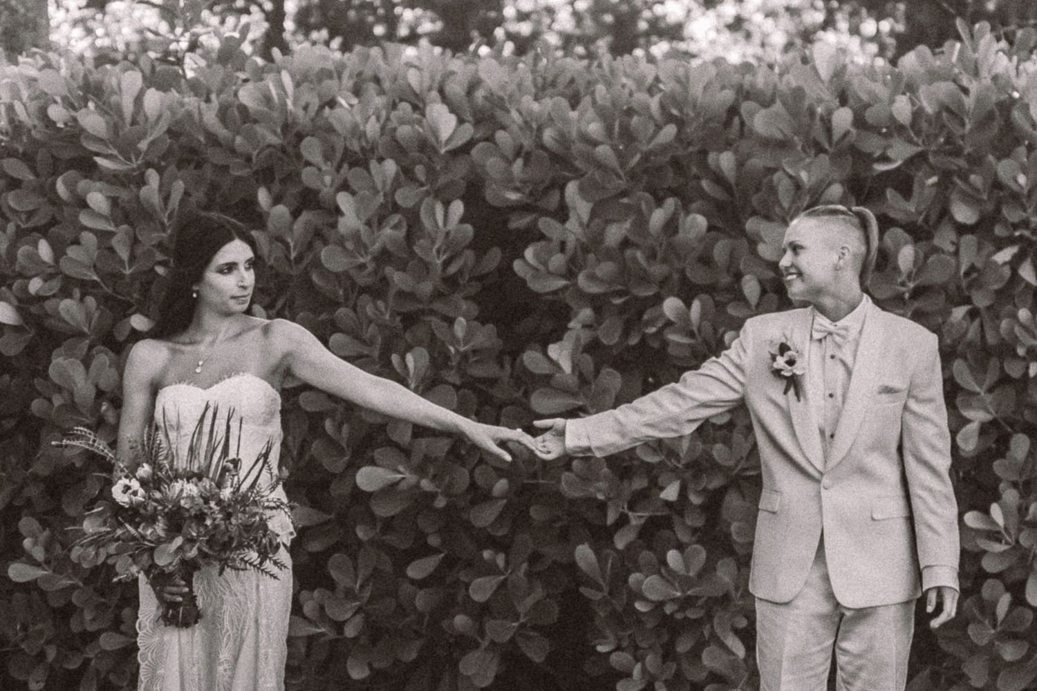 Black and white wedding photo of a couple holding hands against a leafy garden backdrop.