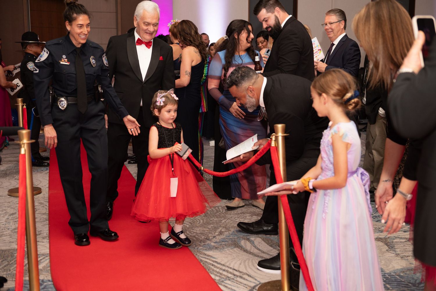 People gather on a red carpet for photos at a formal event with velvet ropes and professional lighting.