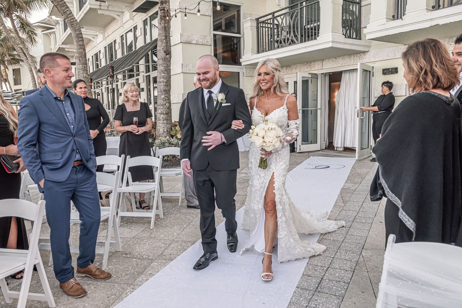 A wedding party walks down a cobblestone street while guests mingle outside a venue on a sunny day.