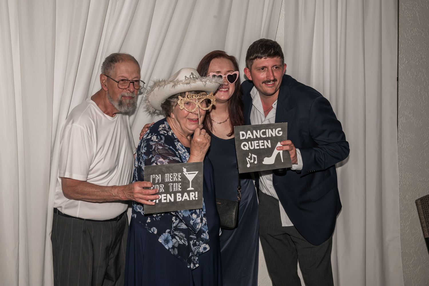 Four people posing at a wedding photo booth, holding fun signs including Dancing Queen, smiling together.