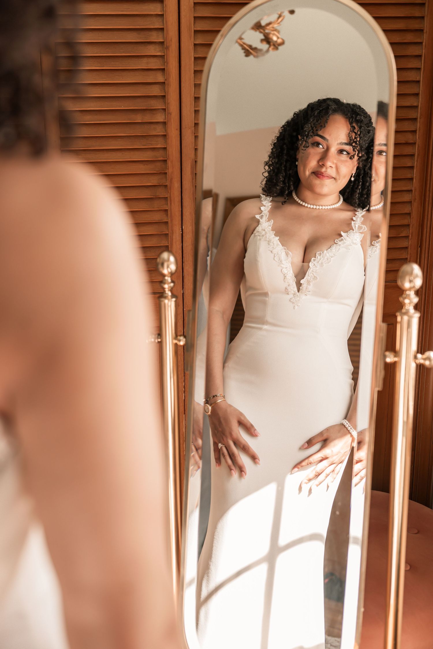 A bride in a white lace gown admires her reflection in a gold-framed full-length mirror.