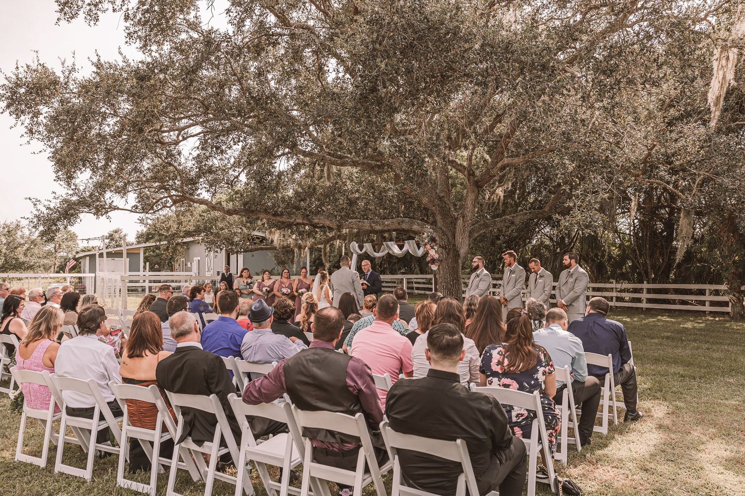 Wedding guests seated in white chairs for an outdoor ceremony under large oak trees at a rustic venue.