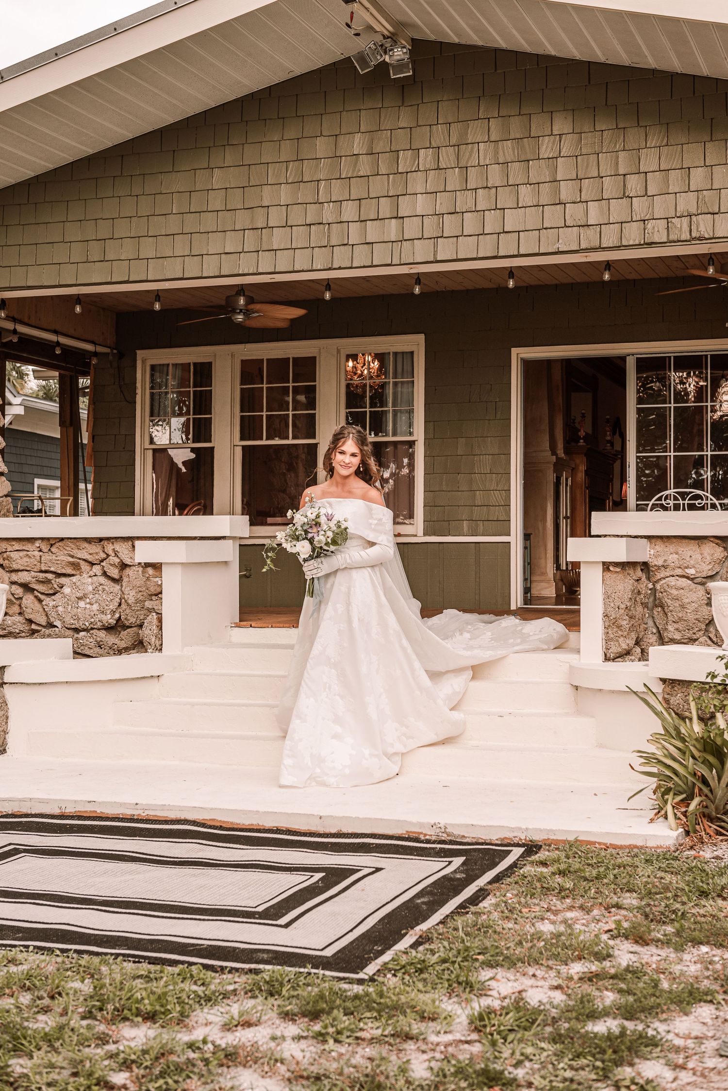 A bride in a flowing white off-shoulder wedding dress stands on the front porch of a craftsman-style home with stone pillars.