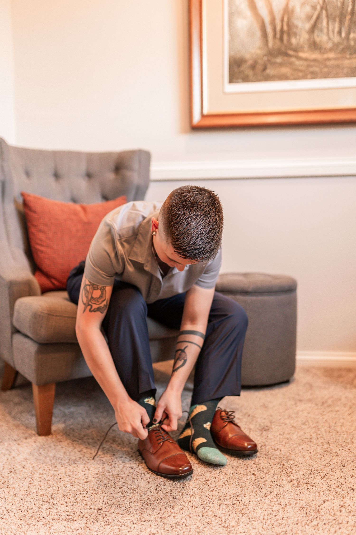 Person sitting on carpet tying brown leather dress shoes in a room with framed artwork on the wall.