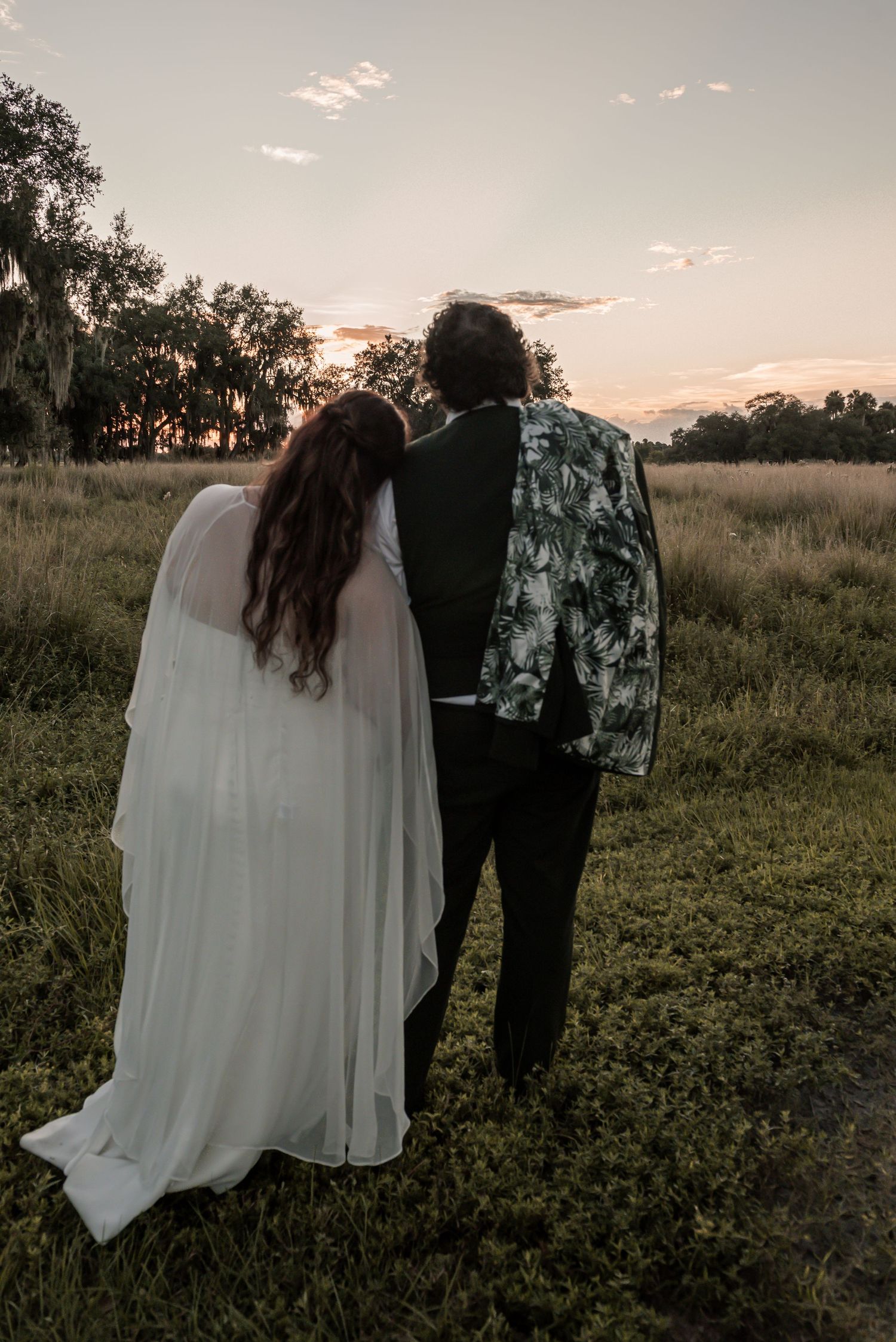 A couple stands in a field at sunset with the bride wearing a flowing white dress against a natural landscape.