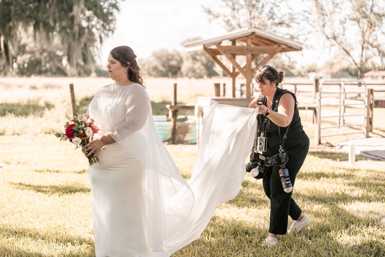 A bride in a white gown holds a bouquet while a photographer adjusts her train outdoors at a rustic venue.