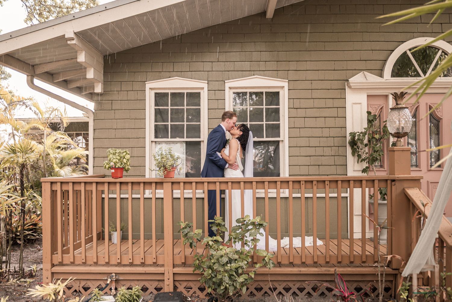 Couple sharing a romantic kiss on a wooden deck of a charming green cottage surrounded by lush plants.