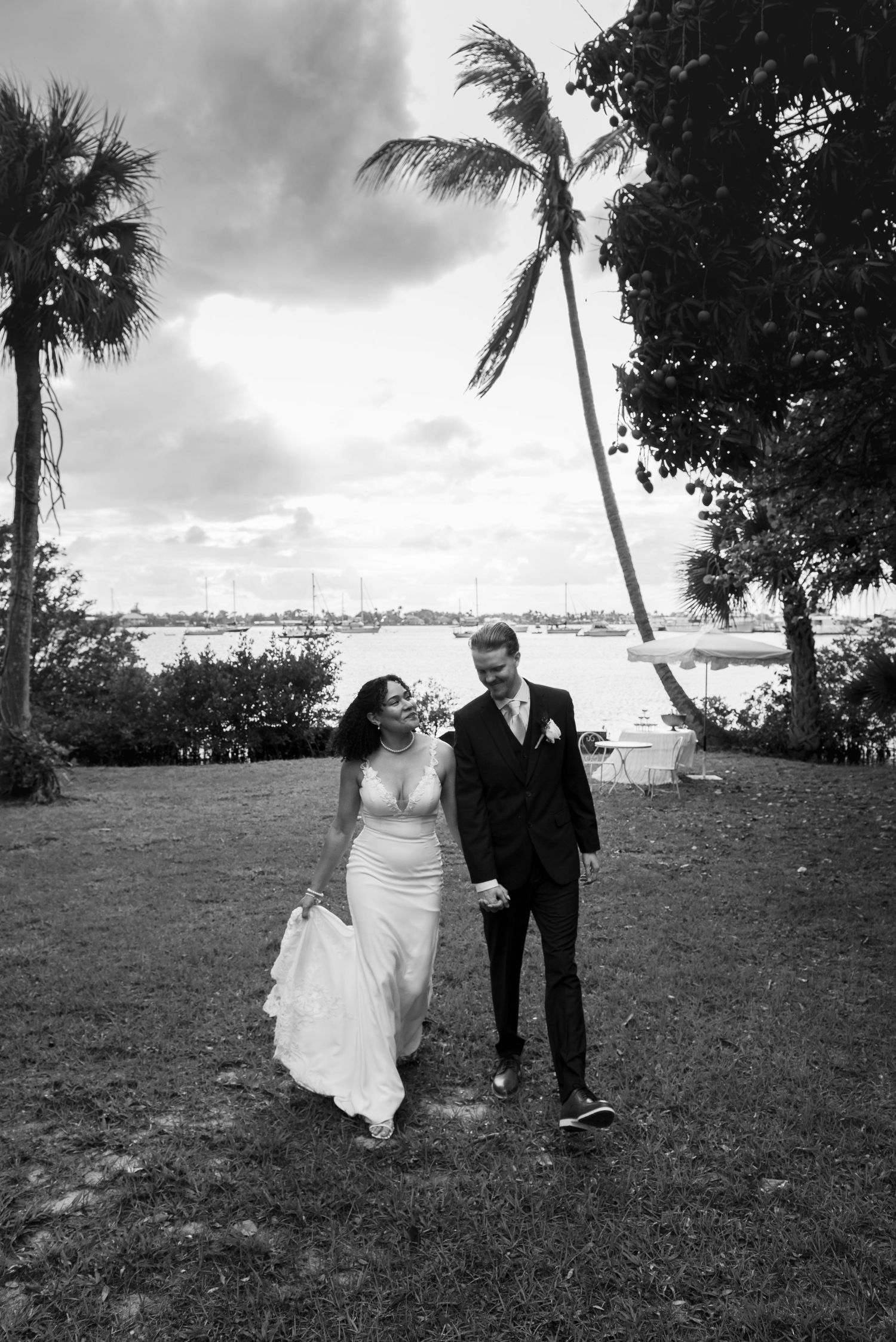 Black and white photo of a bride and groom walking hand in hand on a grassy lawn with palm trees and water in the background.