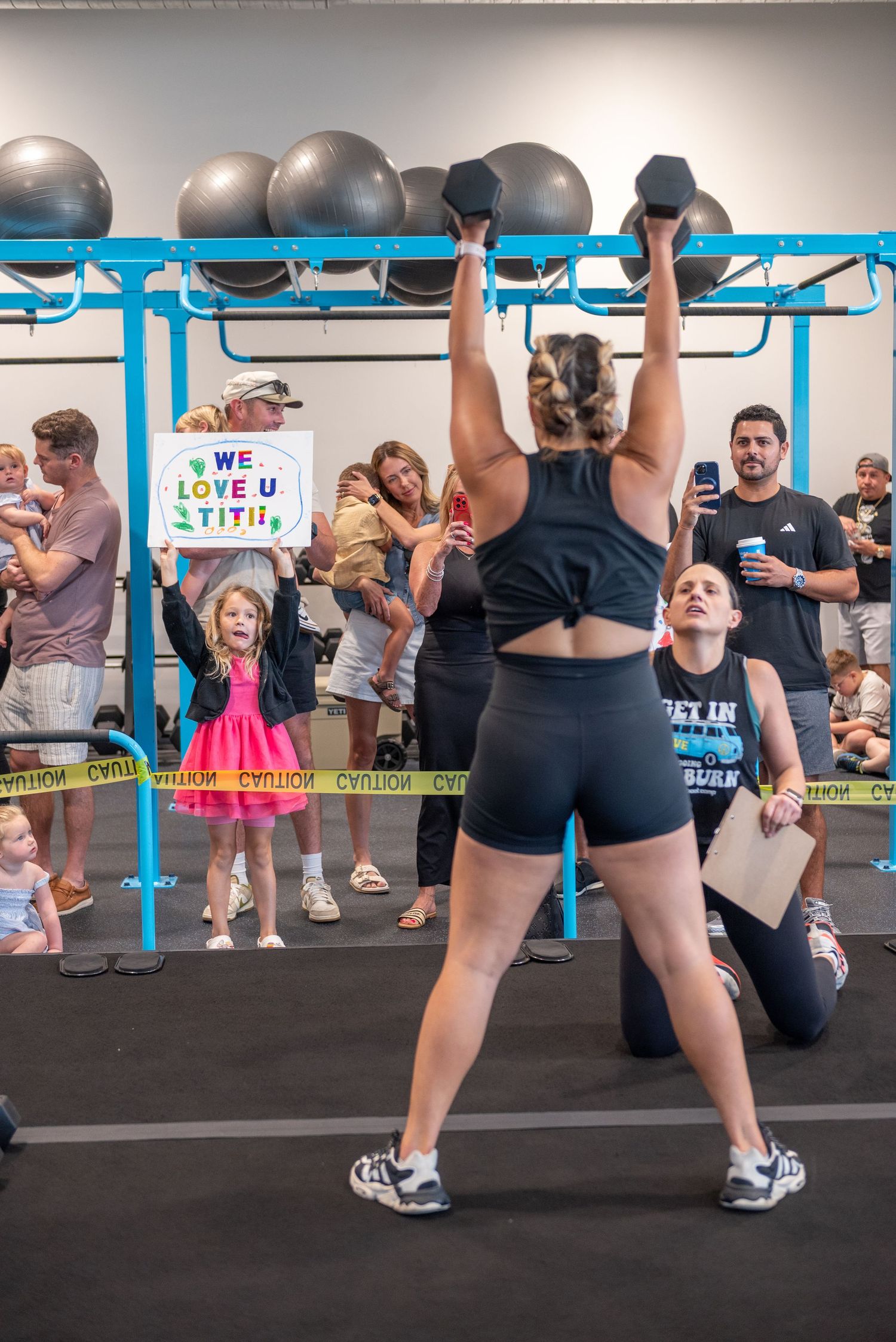Woman lifts dual kettlebells overhead at CrossFit gym competition with cheering crowd.