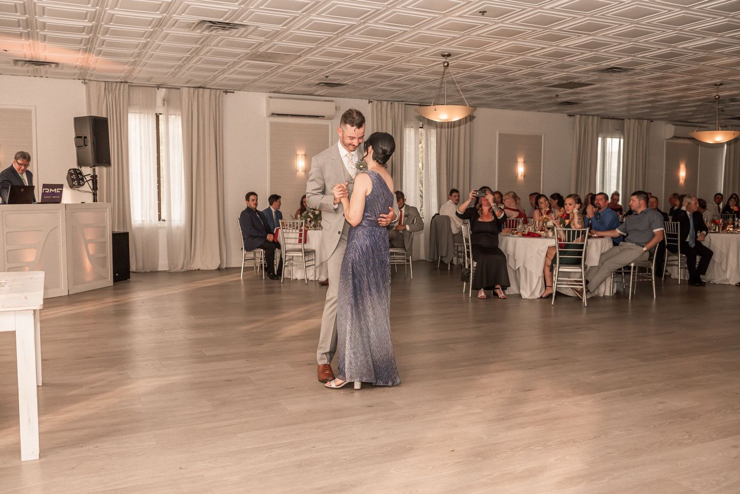 Couple shares their first dance during a wedding reception in an elegant ballroom with guests seated at tables.