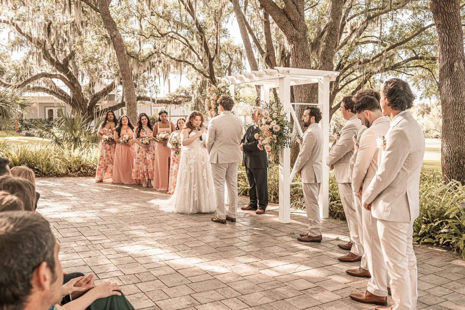 A wedding ceremony takes place outdoors with bridesmaids in pink dresses and groomsmen in beige suits under Spanish moss trees.