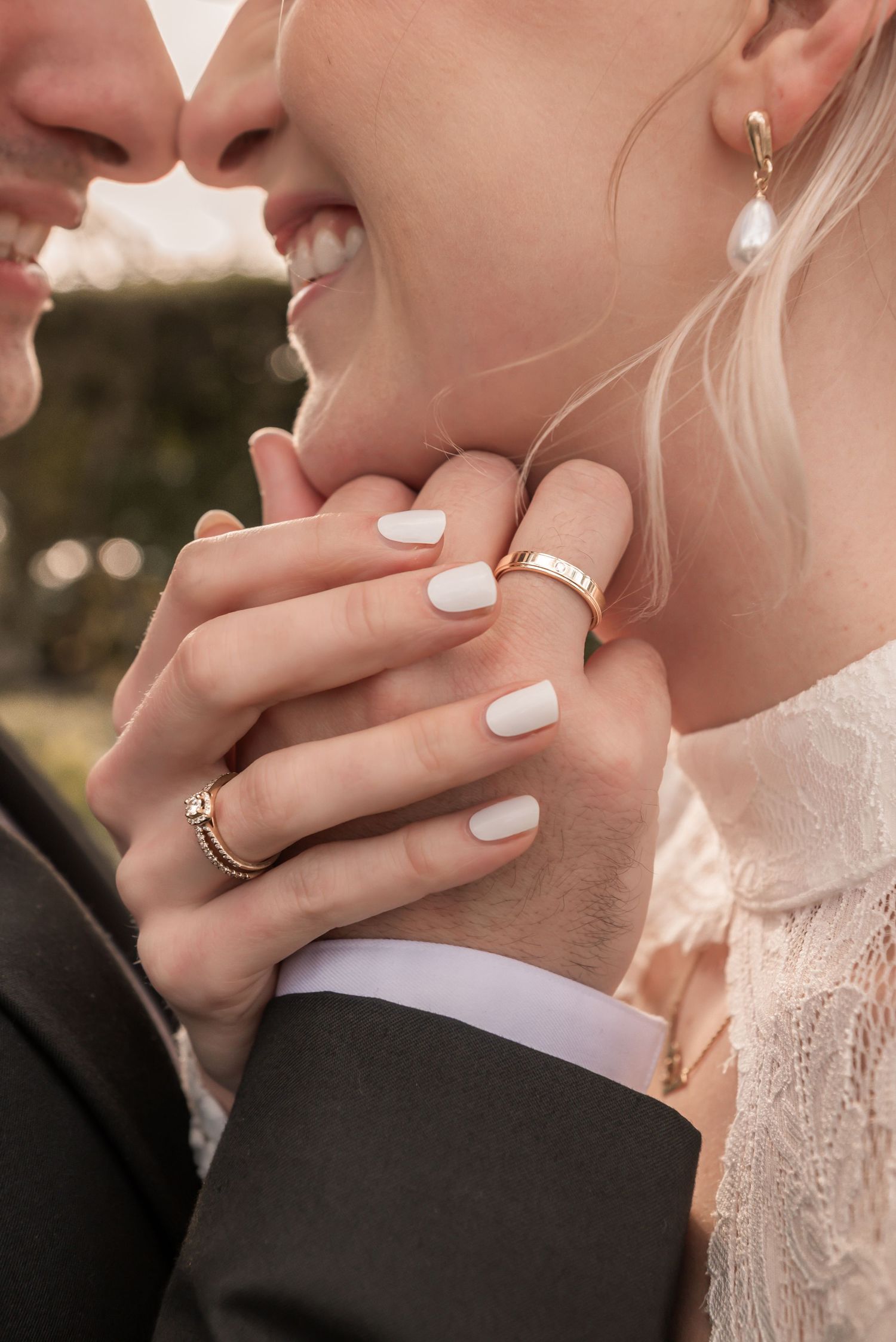 Close-up of wedding couple hands showing gold rings, white manicure, and pearl earring on smiling bride.