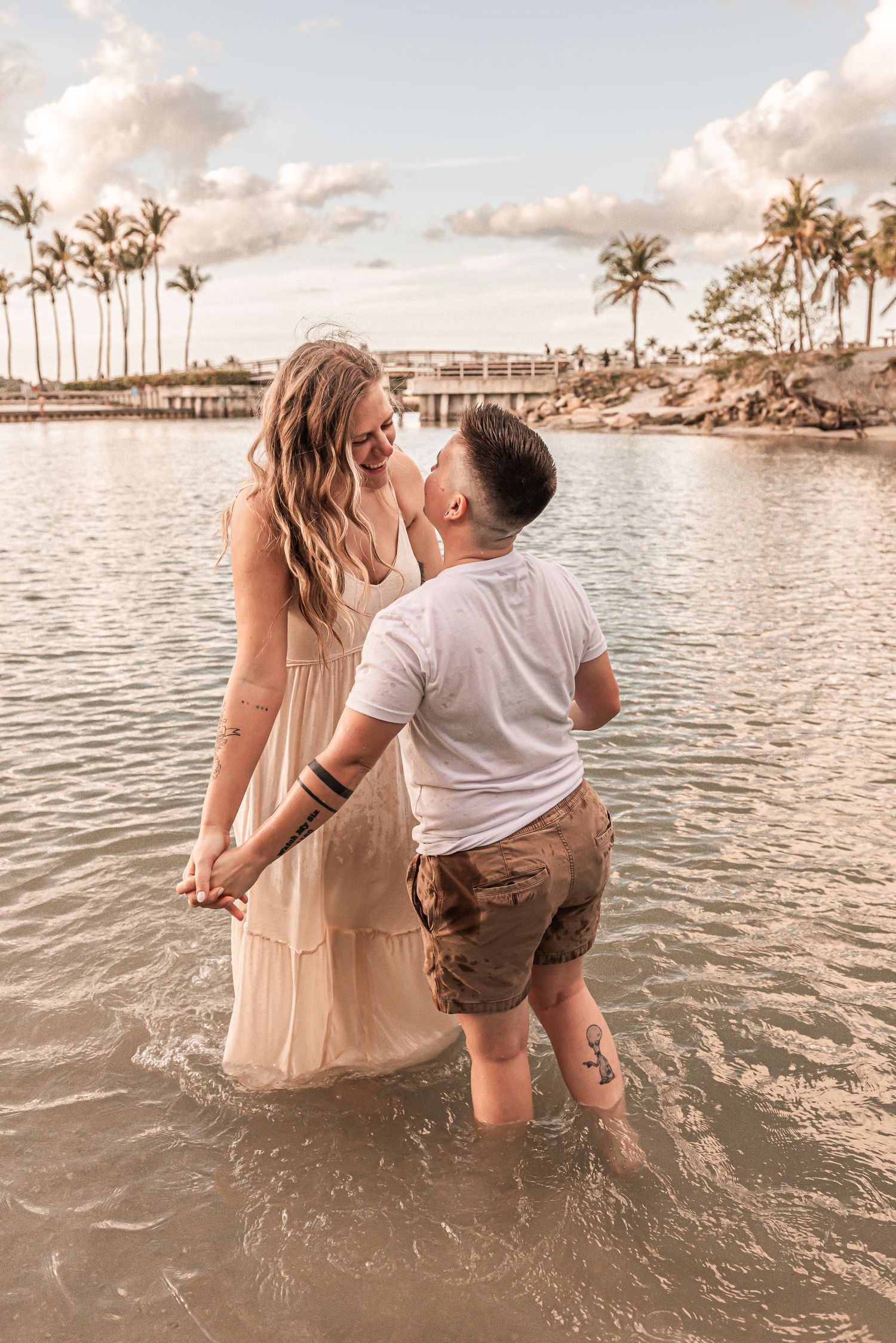 A couple shares a romantic moment while standing in shallow ocean waters at sunset with palm trees in the background.