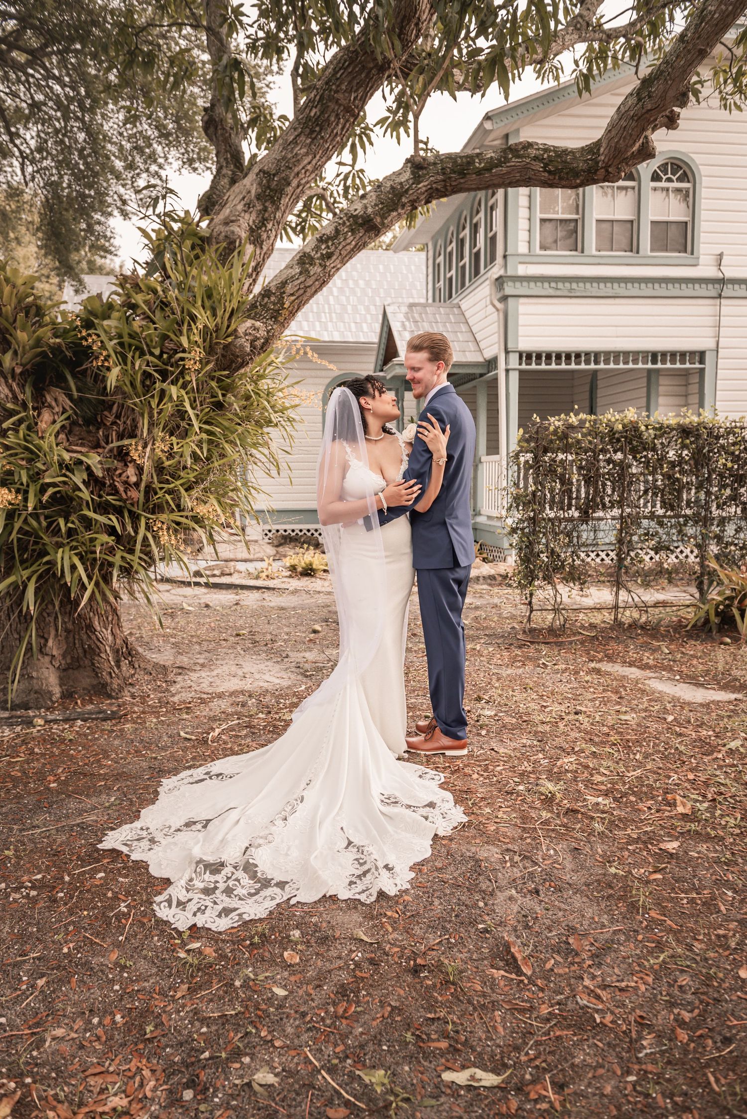 Bride and groom share a romantic embrace in a white mermaid gown and blue suit beneath a large oak tree.