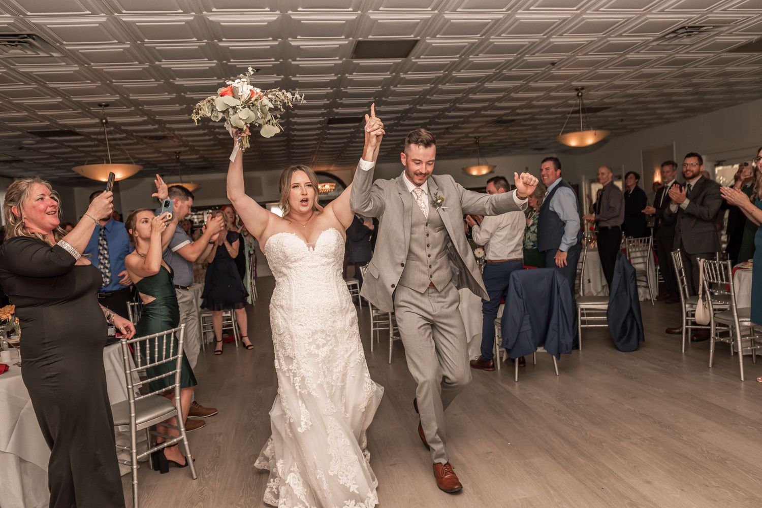 Newlywed couple makes grand entrance into wedding reception as guests cheer at indoor venue with gray decor.