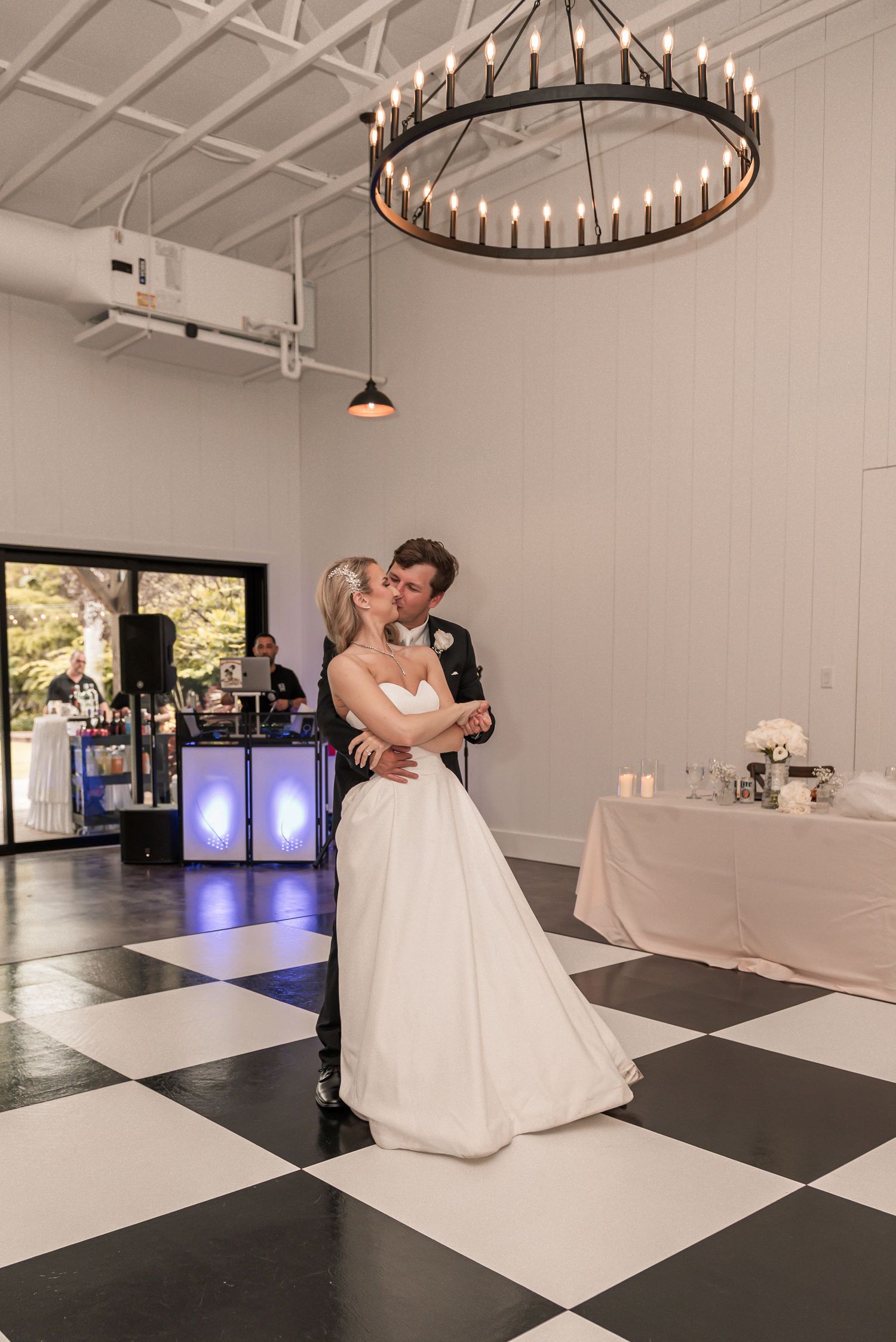 Bride and groom share first dance kiss on checkered floor under chandelier at elegant wedding reception.
