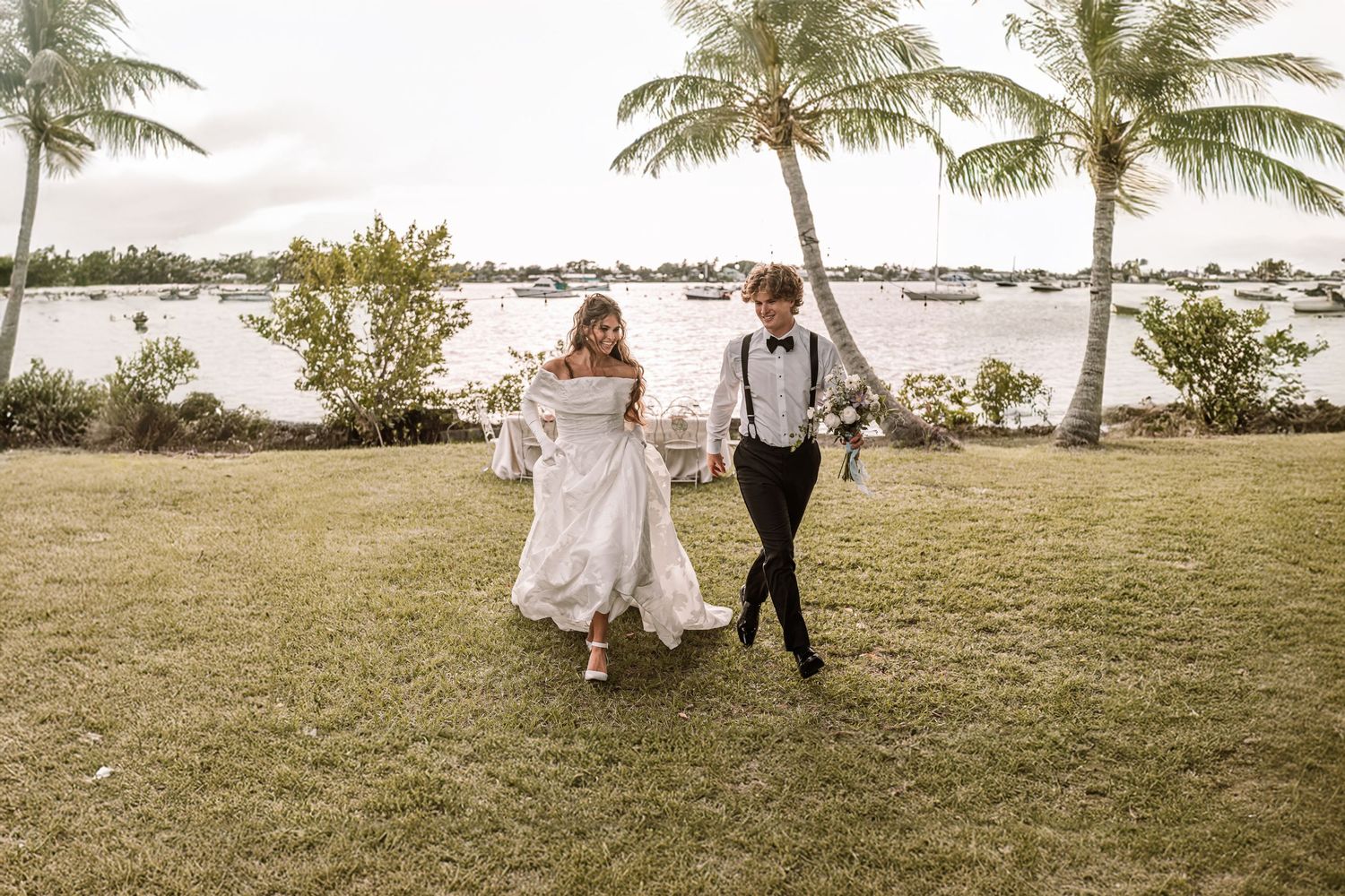 Wedding couple runs joyfully across a waterfront lawn with palm trees and boats in the background.