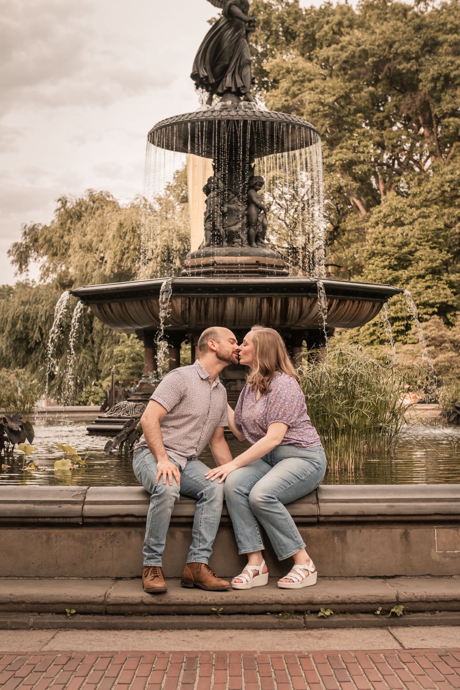 Romantic couple sharing a tender moment while sitting on the edge of an ornate fountain in a park setting.