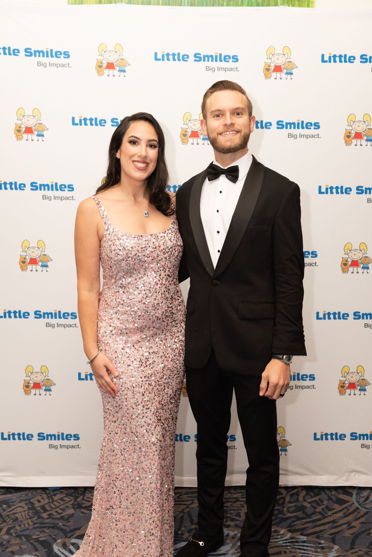 A couple poses together at a formal event with a blue and white branded backdrop at a charity gala.