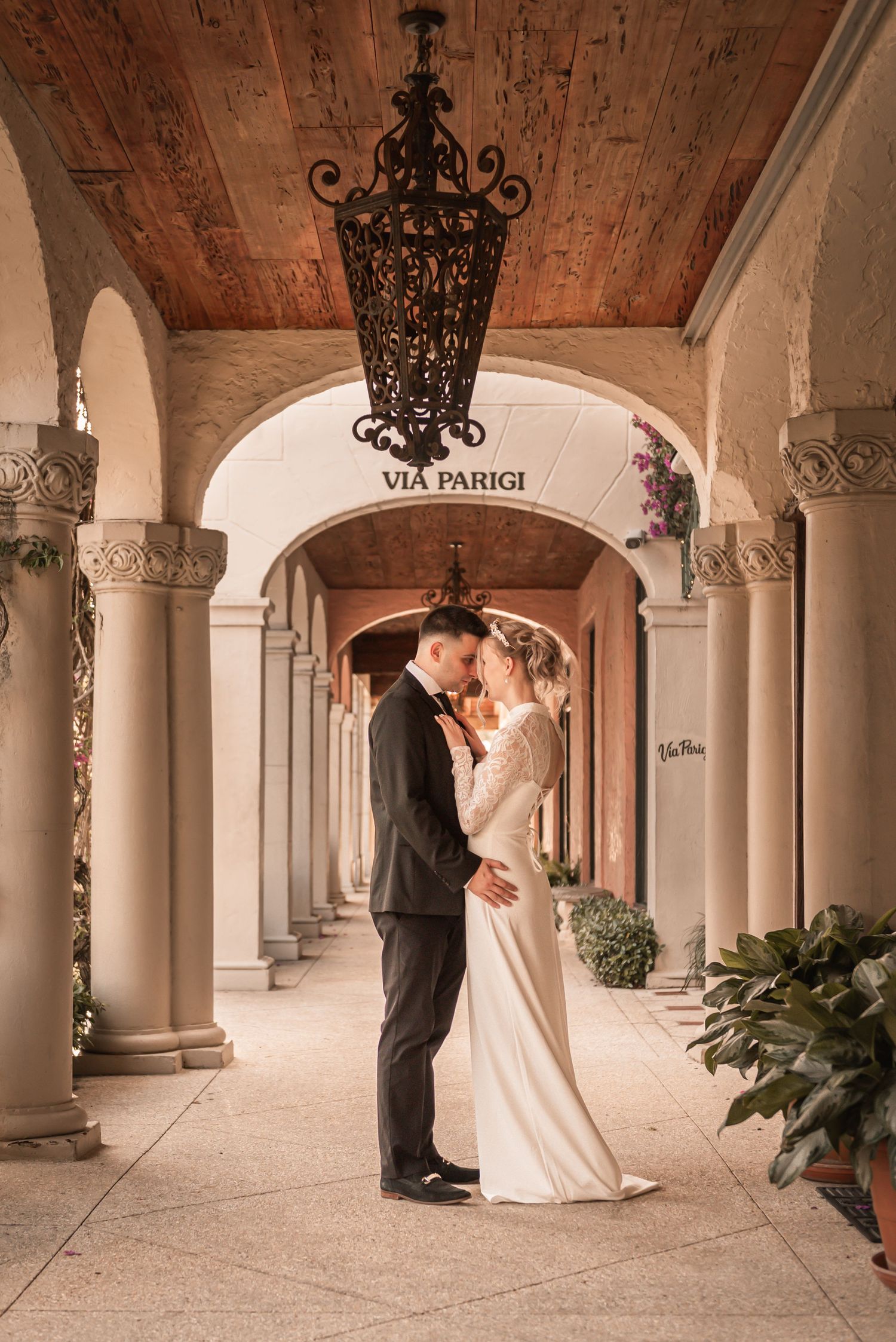 Bride and groom share a romantic moment under elegant arched colonnade at Via Parigi, illuminated by ornate chandelier.