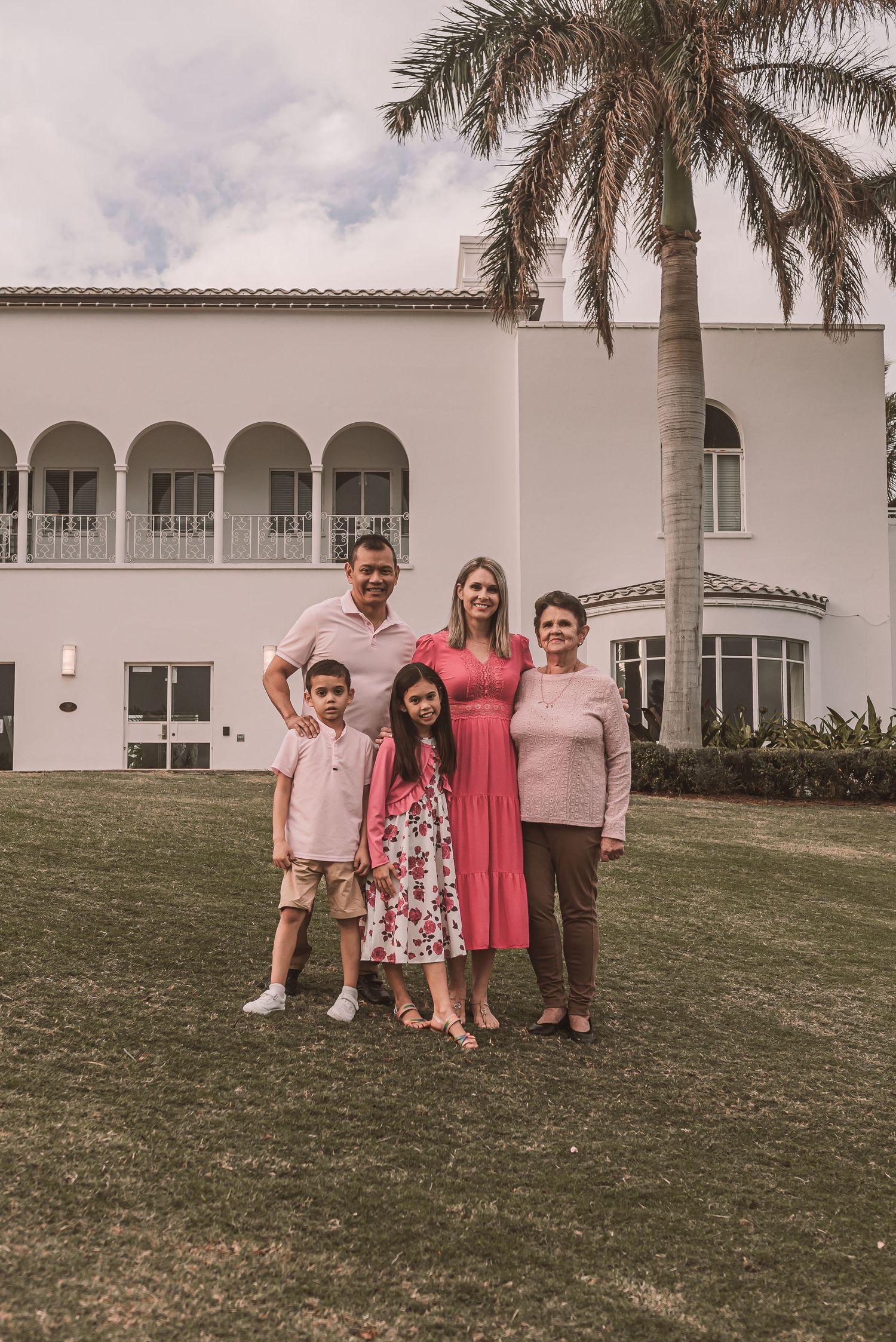 A group stands together on a lawn in front of a white Mediterranean-style mansion with arched windows and tropical palm trees.
