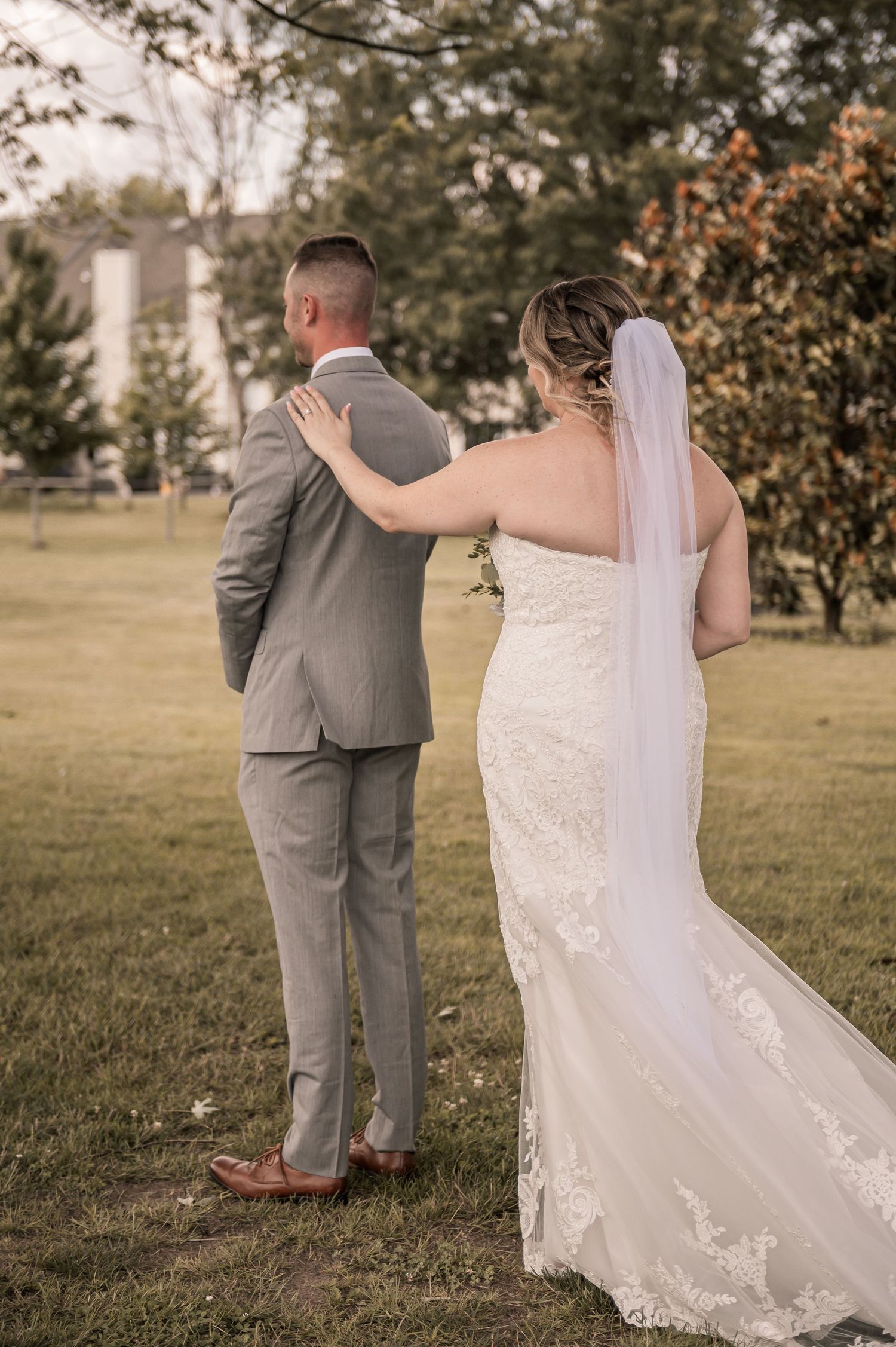A bride in a lace wedding gown touches her groom's shoulder during their first look photo moment in an outdoor garden setting.