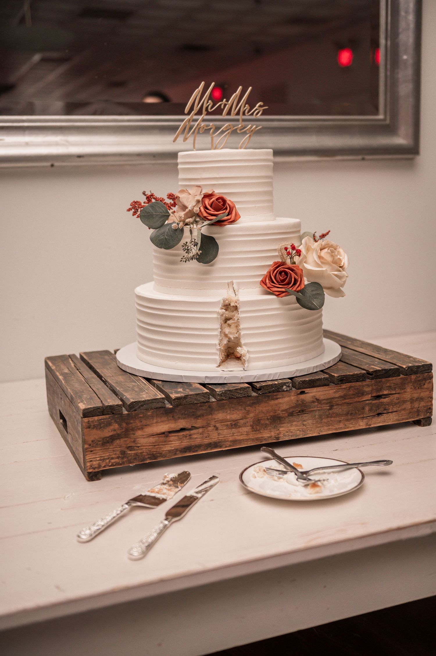Elegant white wedding cake with rose decorations displayed on rustic wooden box with serving utensils.
