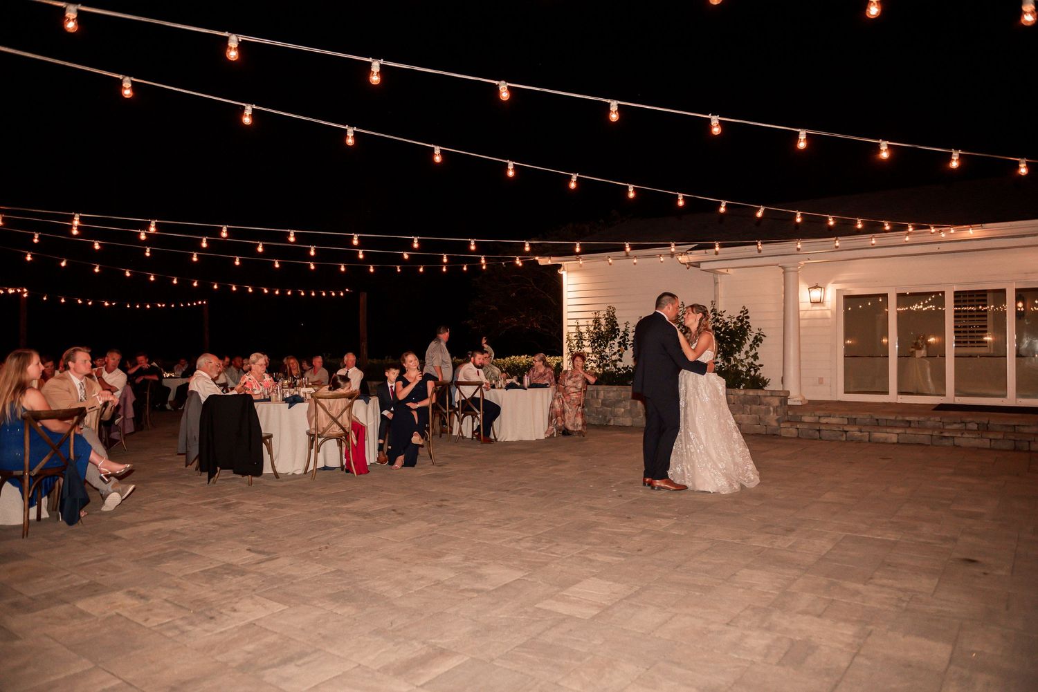 A couple dances under romantic string lights at an outdoor evening wedding reception with guests seated at tables.