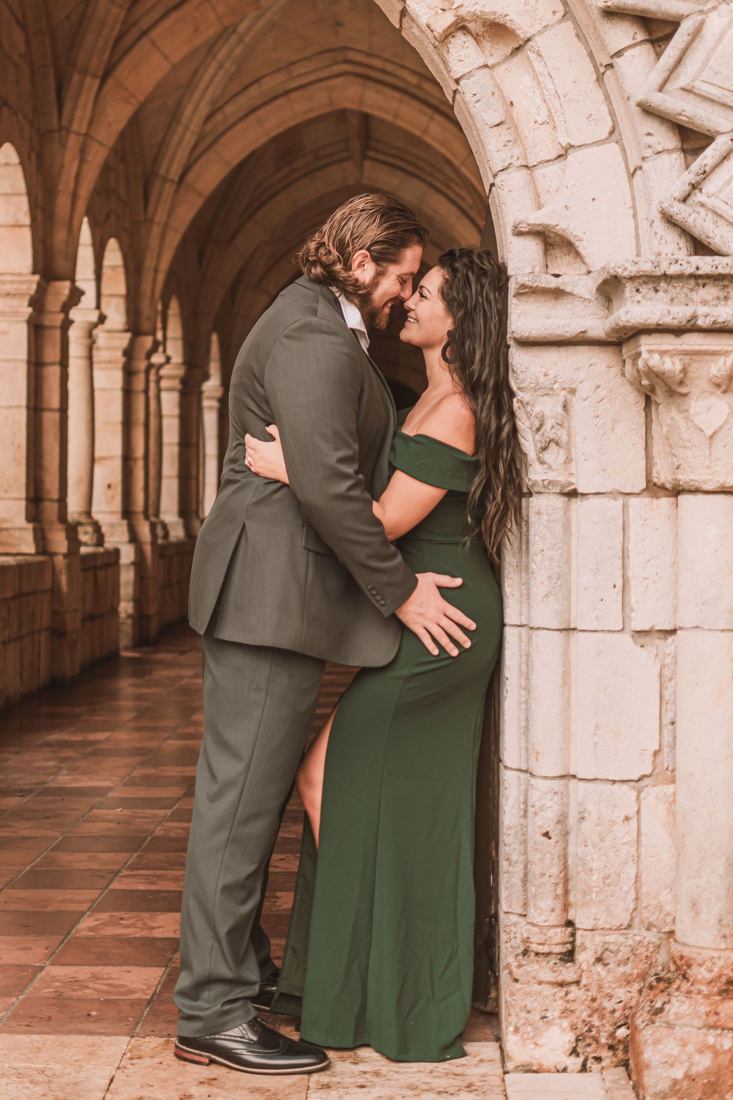 A romantic couple embraces in a stone archway corridor wearing green and grey formal attire.